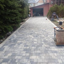 Krzyś - Ułożona kostka brukowa na podjeździe, widoczny pracownik układający kolejne elementy oraz zagęszczarka wibracyjna, w tle garaż i zieleń