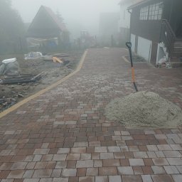 Krzyś - Układanie kostki brukowej na podjeździe w mglisty dzień, widoczna sterta piasku i łopata, w tle budynki mieszkalne i traktor.
