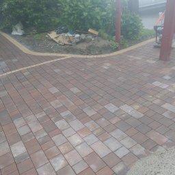 Krzyś - Układanie kostki brukowej w kolorze czerwono-szarym, widoczna krawędź z jasnych kamieni, w tle roślinność i częściowo widoczny samochód dostawczy