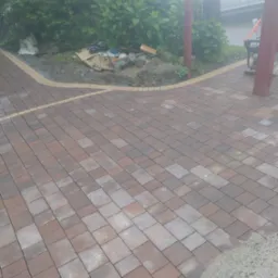 Układanie kostki brukowej w kolorze czerwono-szarym, widoczna krawędź z jasnych kamieni, w tle roślinność i częściowo widoczny samochód dostawczy