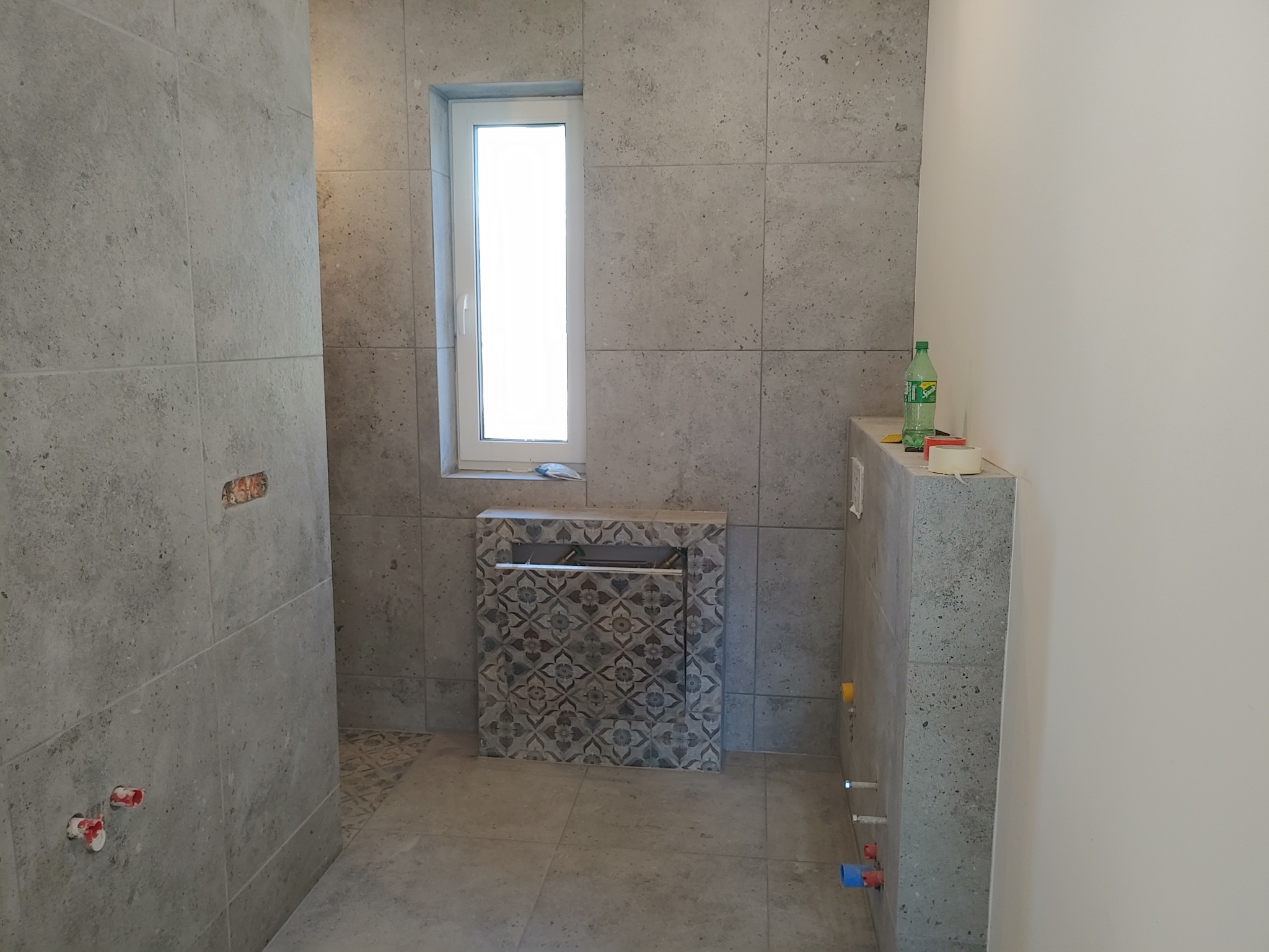 Wnętrze łazienki w trakcie wykańczania, widoczne szare płytki na ścianach i podłodze, zabudowa stelaża podtynkowego WC z dekoracyjnymi płytkami, okno oraz instalacje wodno-kanalizacyjne.