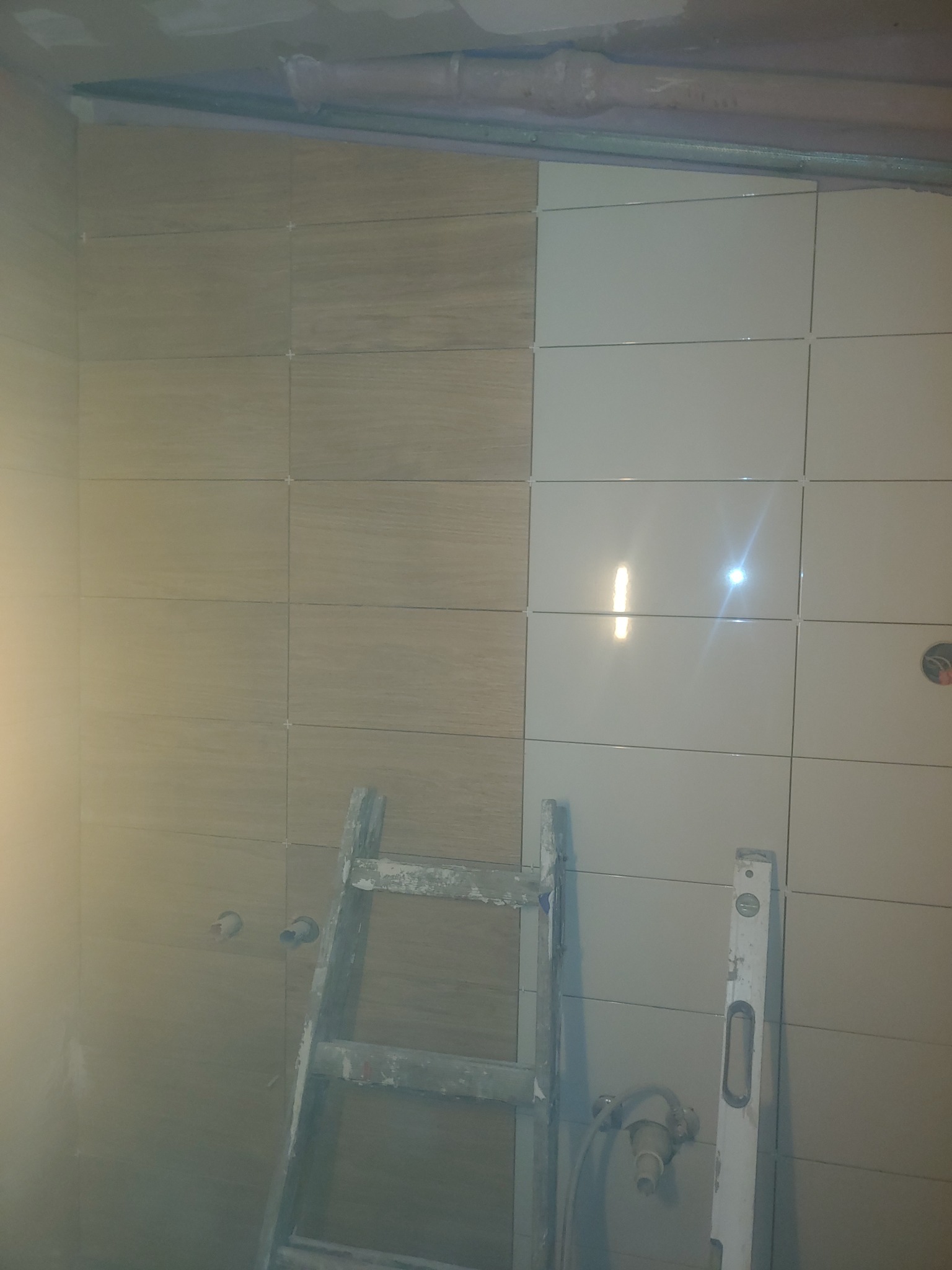 Częściowo wykończona łazienka z dwoma rodzajami płytek, drabina i poziomica oparta o ścianę, widoczne instalacje wodne.