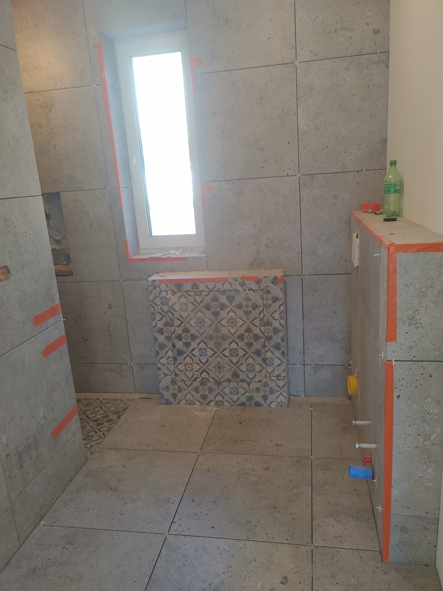 Łazienka w trakcie wykańczania: szare, kwadratowe płytki na ścianach i podłodze, okno z pomarańczową taśmą, zabudowa WC z dekoracyjnymi płytkami i butelka wody na górze, widoczne elementy...