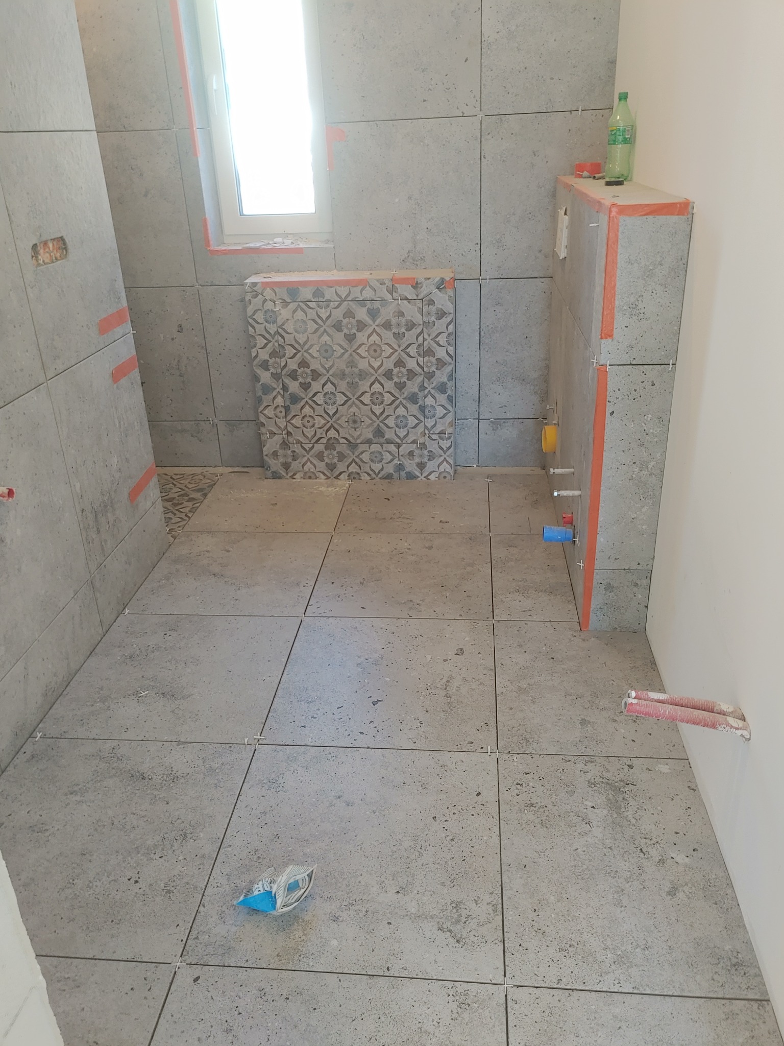 Szare płytki podłogowe i ścienne w trakcie montażu w łazience, widoczne krzyżyki dystansowe i elementy instalacji hydraulicznej, butelka z płynem na obudowie stelaża podtynkowego.