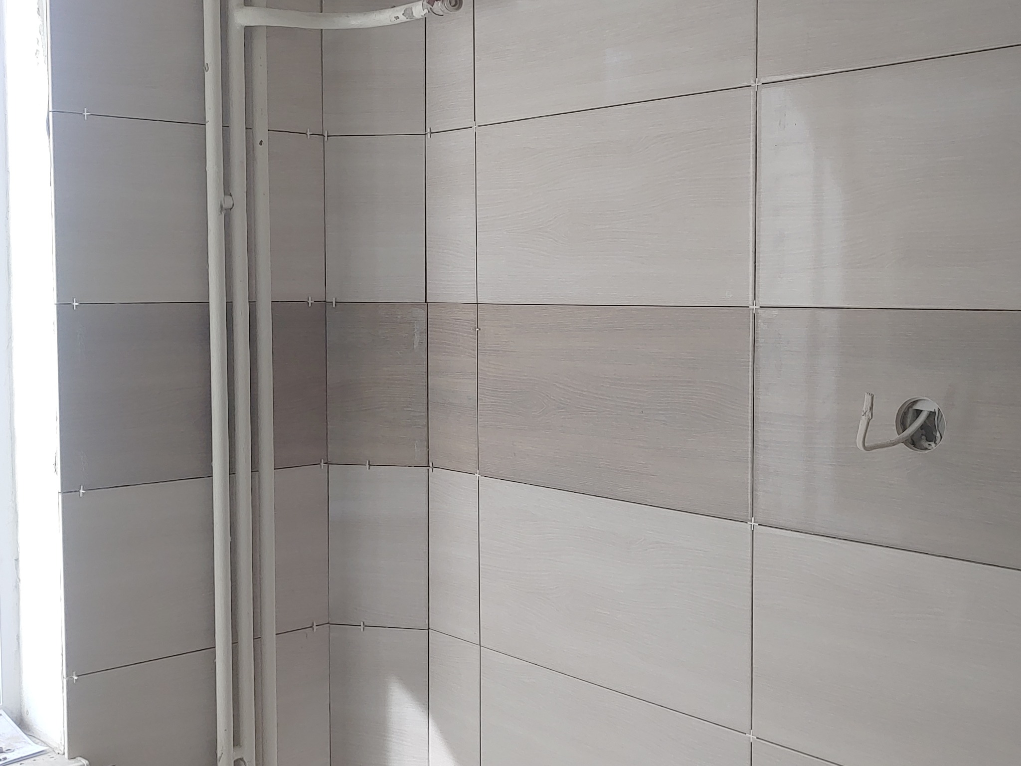 Ściana w trakcie wykańczania z ułożonymi płytkami ceramicznymi w odcieniach szarości i beżu z imitacją drewna, widoczne krzyżyki dystansowe, rury instalacji grzewczej i otwór na gniazdko elektryczne.