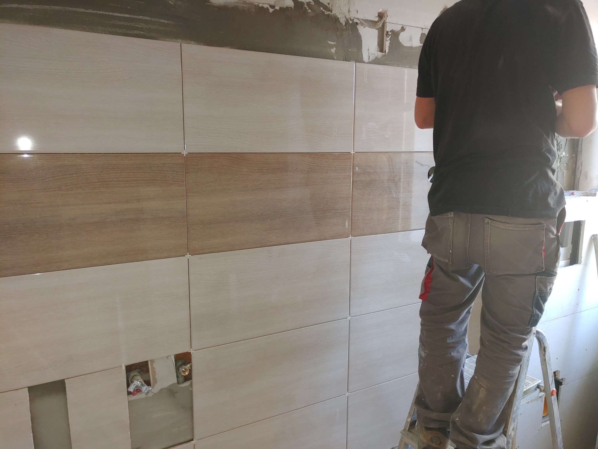 Układanie płytek imitujących drewno na ścianie, widoczne krzyżyki dystansowe i otwory na instalacje wodne, pracownik na drabinie w tle.