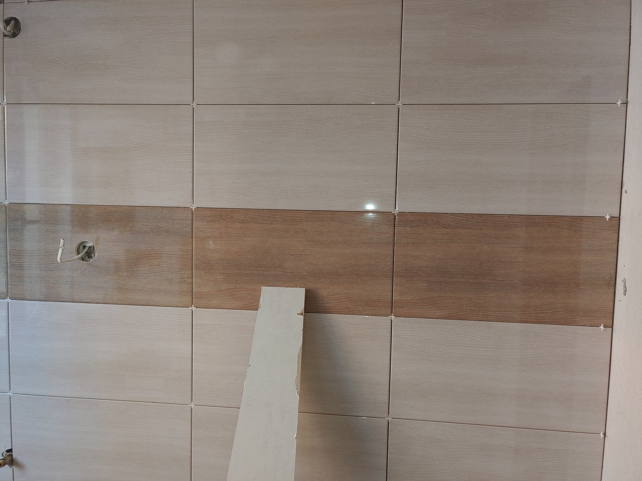 Ściana w trakcie wykańczania z ułożonymi płytkami imitującymi drewno w dwóch odcieniach, z widocznymi krzyżykami dystansowymi i otworem na instalację.