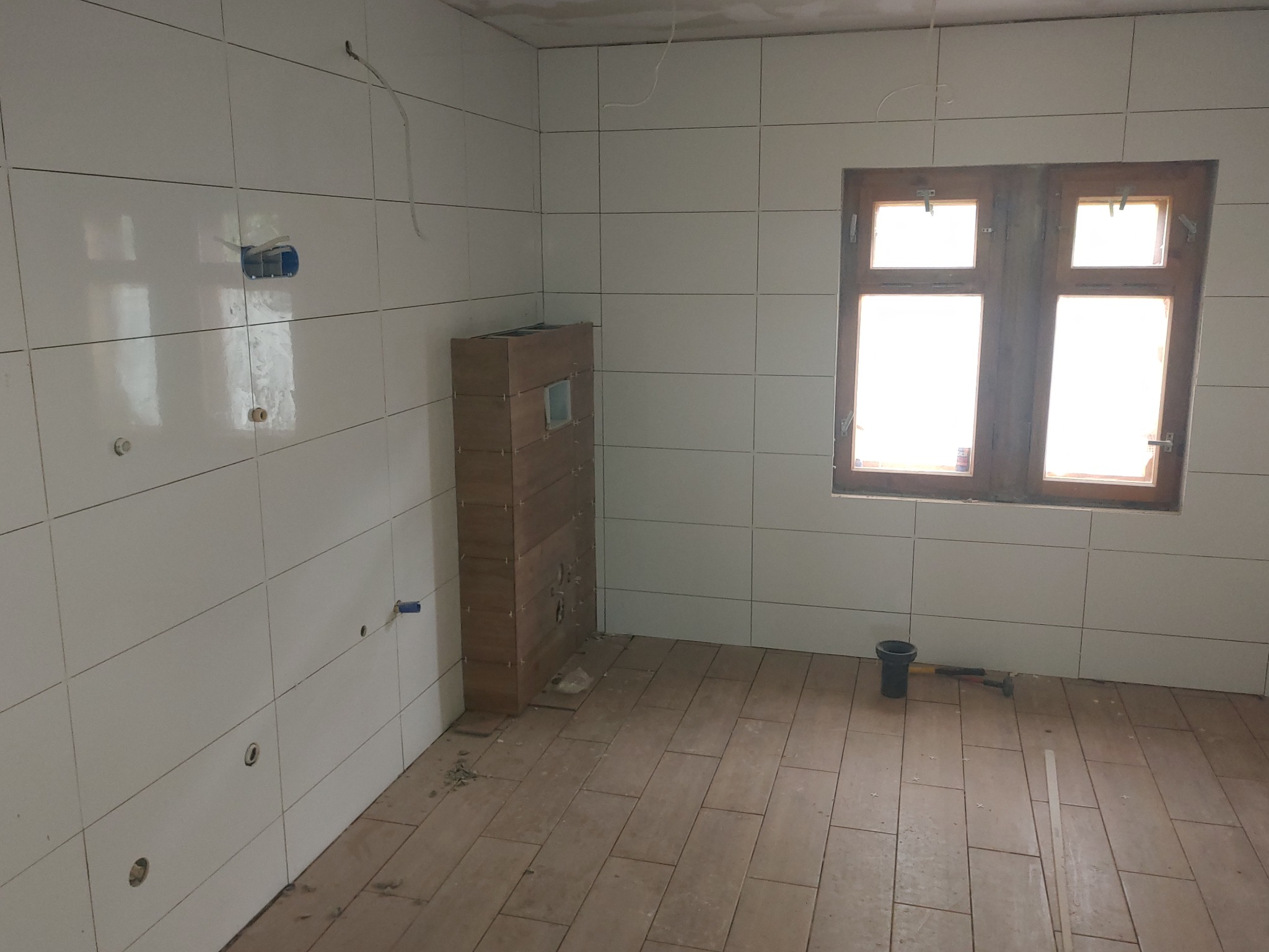 Wnętrze łazienki w trakcie wykańczania: białe płytki ścienne z widocznymi otworami na instalacje, drewnopodobne płytki podłogowe, zabudowa stelaża WC, drewniane okno z jasnym światłem dziennym...