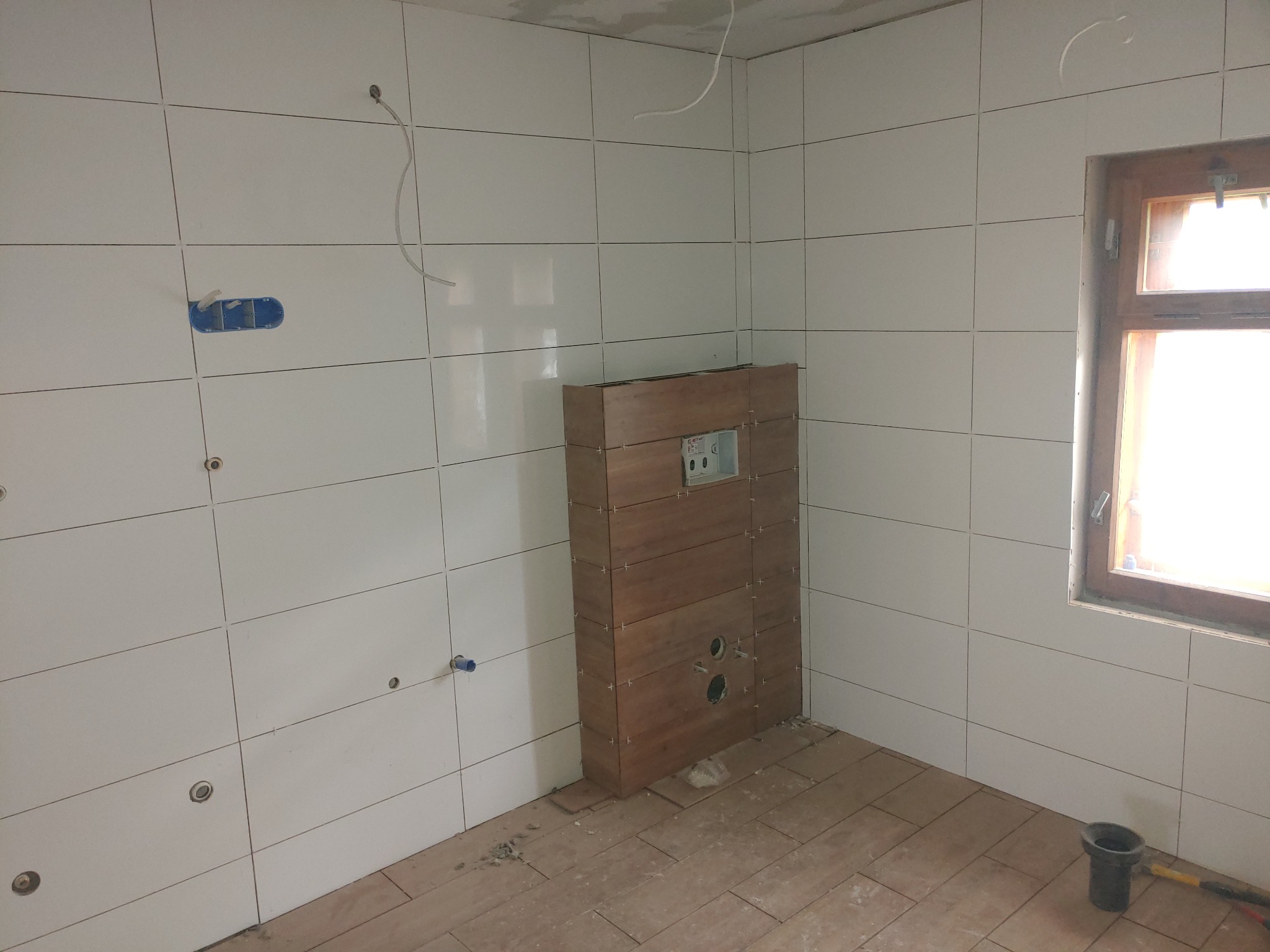 Białe płytki ścienne z widocznymi otworami na instalacje elektryczne i wodne, zabudowa stelaża WC z drewnopodobnych paneli, brudna podłoga z ułożonych płytek, okno w drewnianej ramie, etap prac...