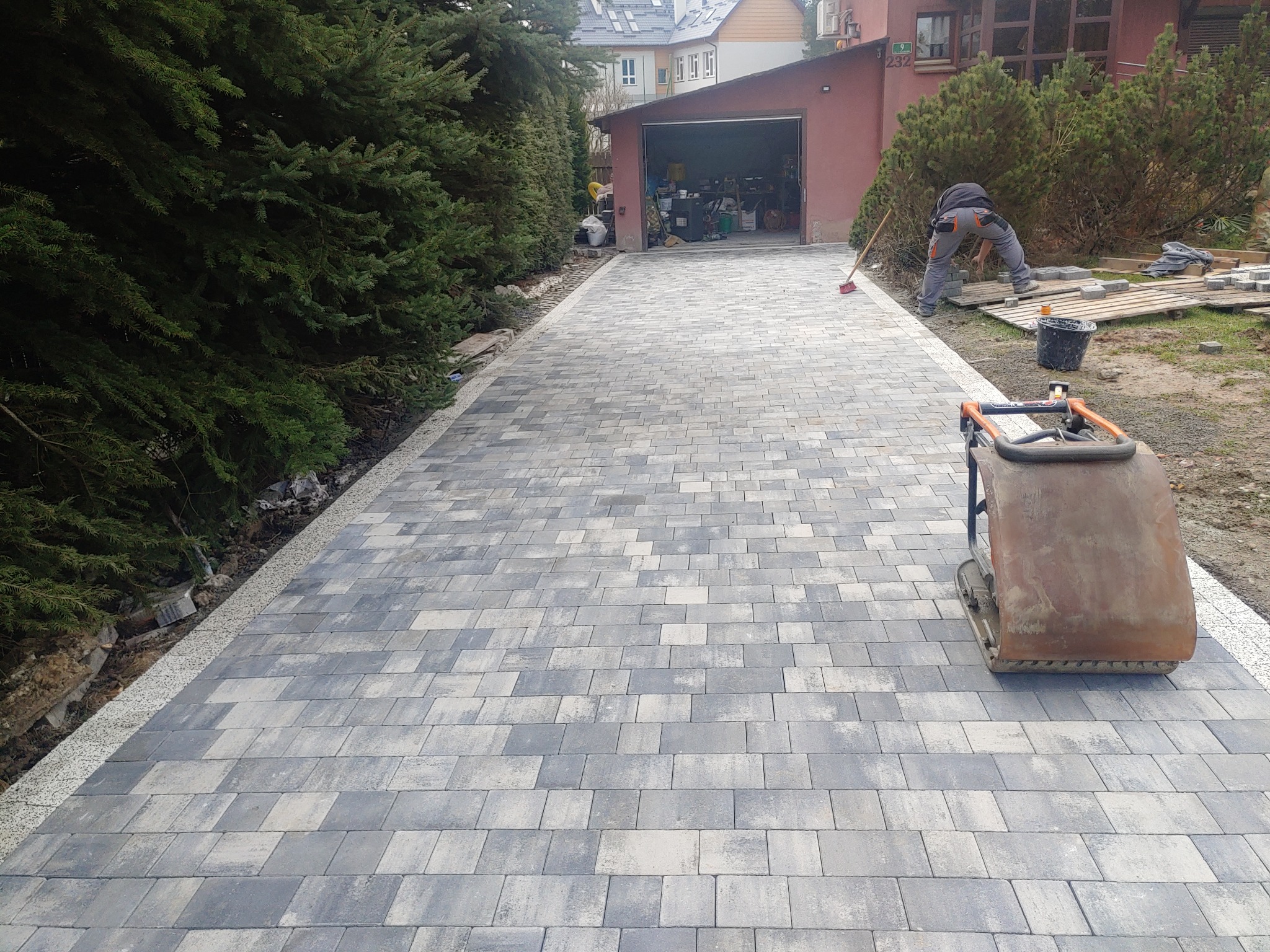 Ułożona kostka brukowa na podjeździe, widoczny pracownik układający kolejne elementy oraz zagęszczarka wibracyjna, w tle garaż i zieleń