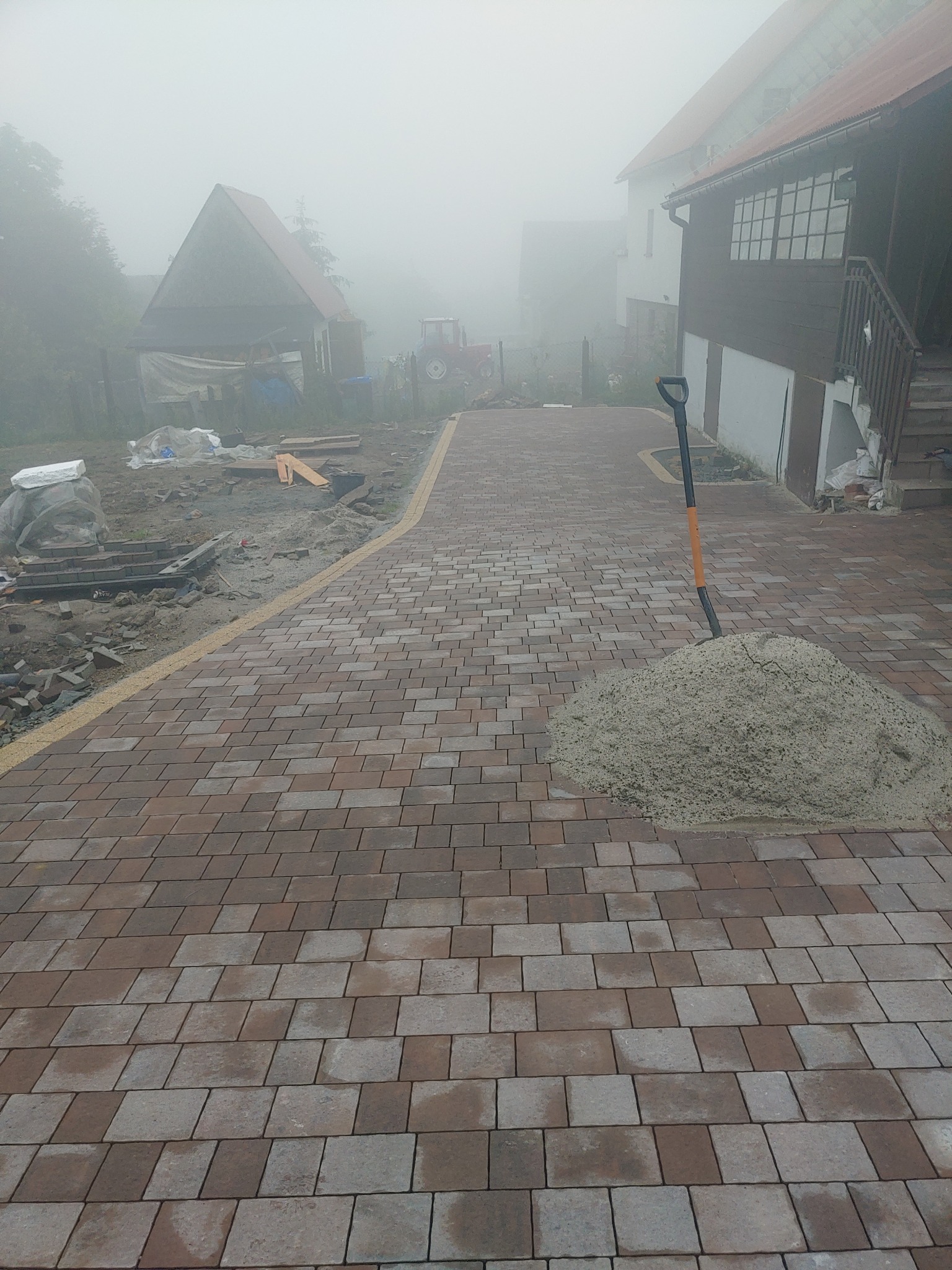 Układanie kostki brukowej na podjeździe w mglisty dzień, widoczna sterta piasku i łopata, w tle budynki mieszkalne i traktor.