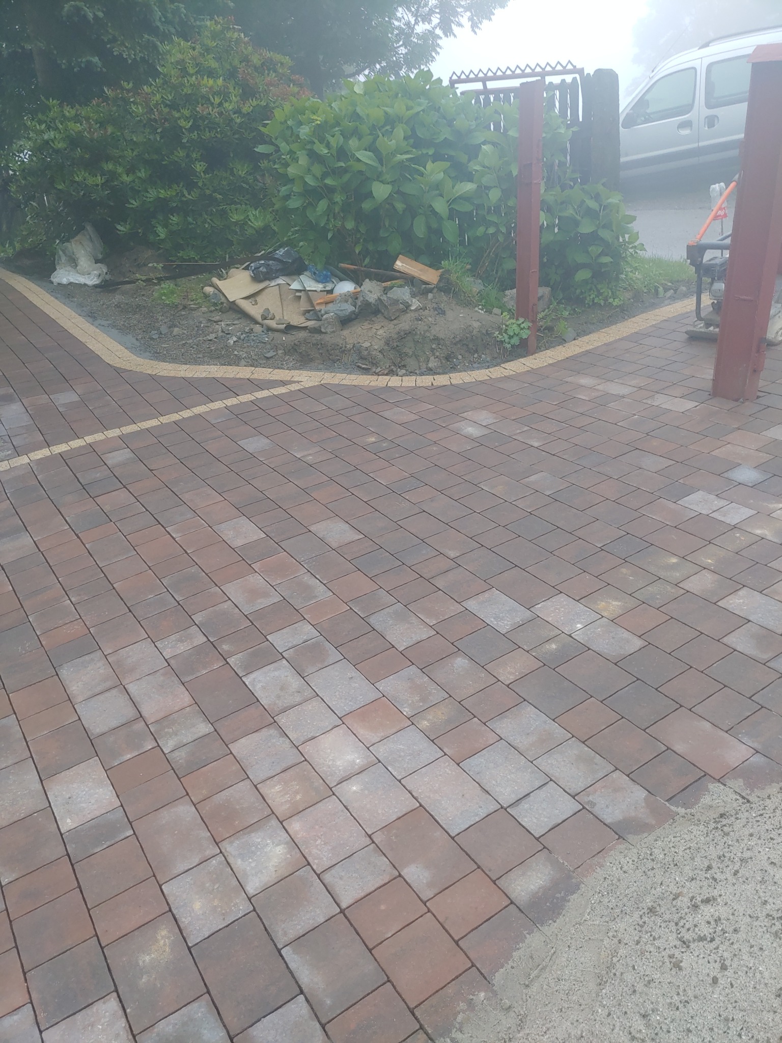 Układanie kostki brukowej w kolorze czerwono-szarym, widoczna krawędź z jasnych kamieni, w tle roślinność i częściowo widoczny samochód dostawczy