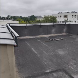 Mobilny Spawacz Jan Bednarczyk Professional Spaw 303 - Szara balustrada na dachu budynku z widokiem na zieleń i inne budynki. Dach pokryty papą, fragmenty drewnianych elementów. Pochmurne niebo w tle.