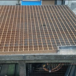 Mobilny Spawacz Jan Bednarczyk Professional Spaw 303 - Metalowa krata z kwadratowymi oczkami, częściowo obramowana, leży na stole warsztatowym, obok narzędzia i rękawice robocze. Widoczny spaw na obramowaniu.