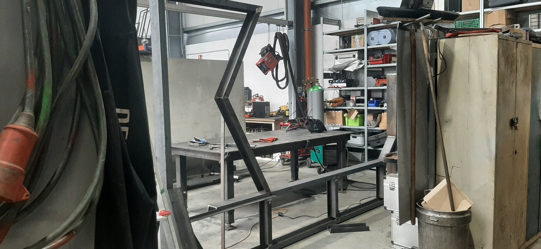 W warsztacie metalowym, konstrukcja z profili stalowych w trakcie spawania. Widoczne narzędzia, stół spawalniczy, butla z gazem i regał z akcesoriami.