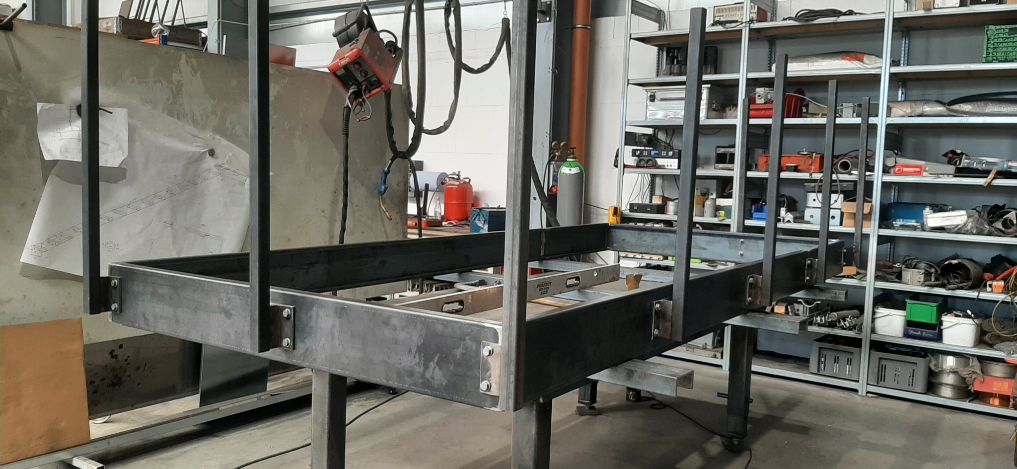 Konstrukcja metalowa z profili, spawana, w warsztacie. Widoczny spawacz, butle z gazem i regał z narzędziami w tle. Element konstrukcyjny, prawdopodobnie schody lub balustrada.