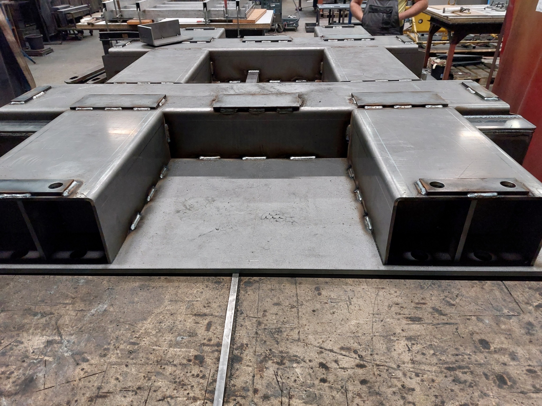 Spawana konstrukcja stalowa z profili zamkniętych, widoczne spawy i otwory, surowy metal, na stole warsztatowym z śladami użytkowania, w tle warsztat.