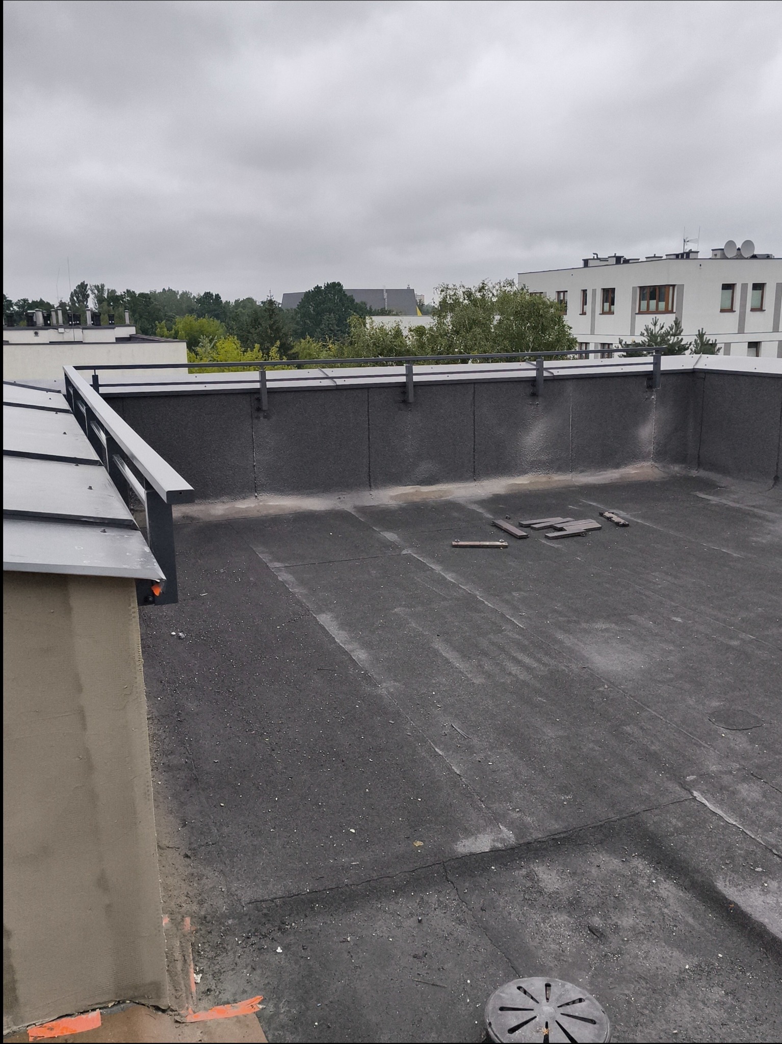 Szara balustrada na dachu budynku z widokiem na zieleń i inne budynki. Dach pokryty papą, fragmenty drewnianych elementów. Pochmurne niebo w tle.