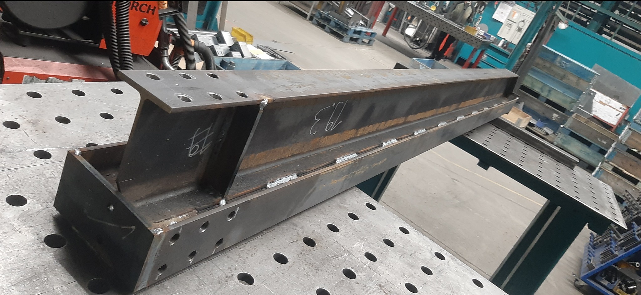 Spawana konstrukcja stalowa z otworami na stole spawalniczym. Widoczne ślady spawania, numery markerem. Tło warsztatowe, elementy wyposażenia.