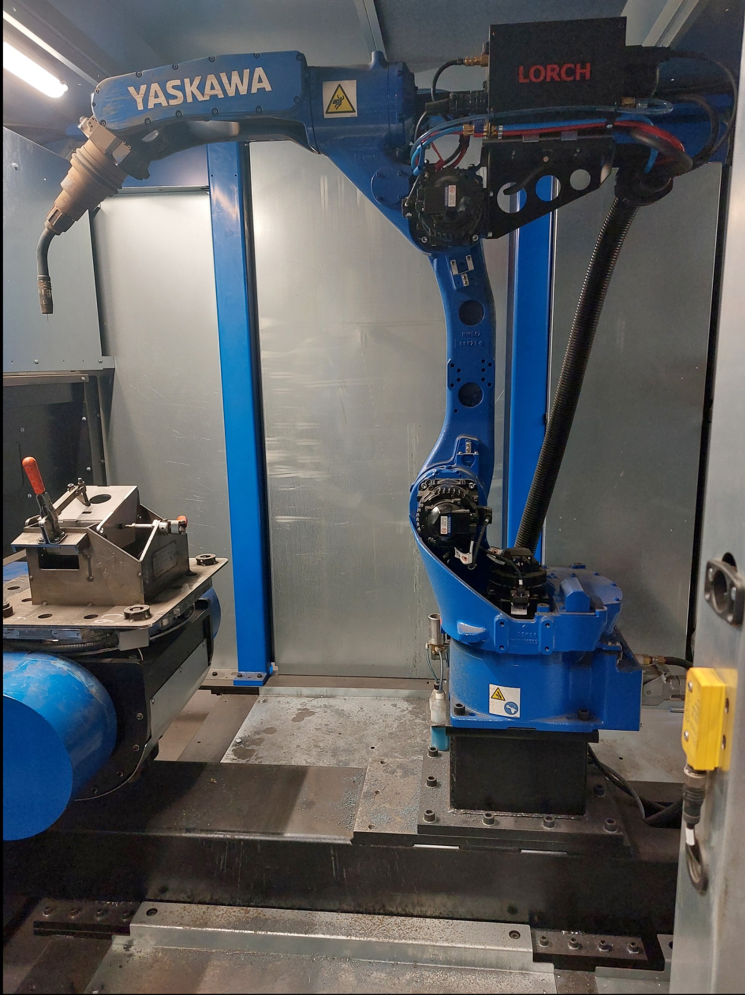 Niebieski robot spawalniczy Yaskawa z osprzętem Lorch w szarej kabinie, uchwyt mocujący element obrabiany. Automat do spawania metali w procesie produkcyjnym.