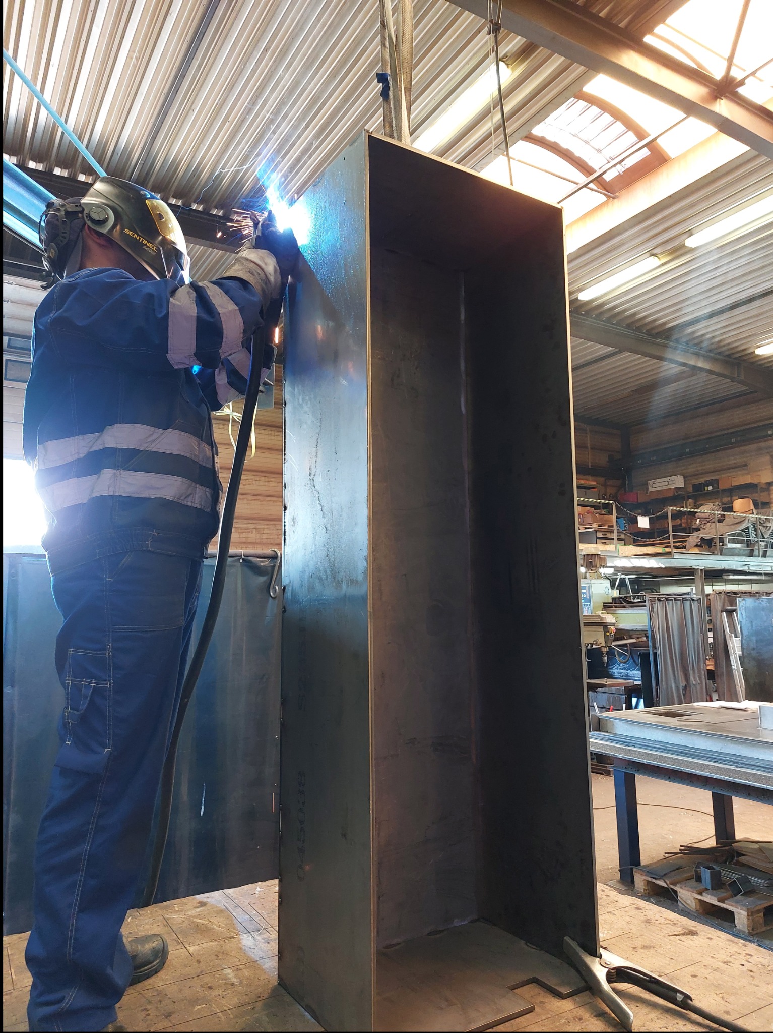 Pracownik w kombinezonie spawalniczym łączy elementy metalowe w hali produkcyjnej. Widoczne iskry i spawarka. W tle metalowe konstrukcje i oświetlenie przemysłowe.