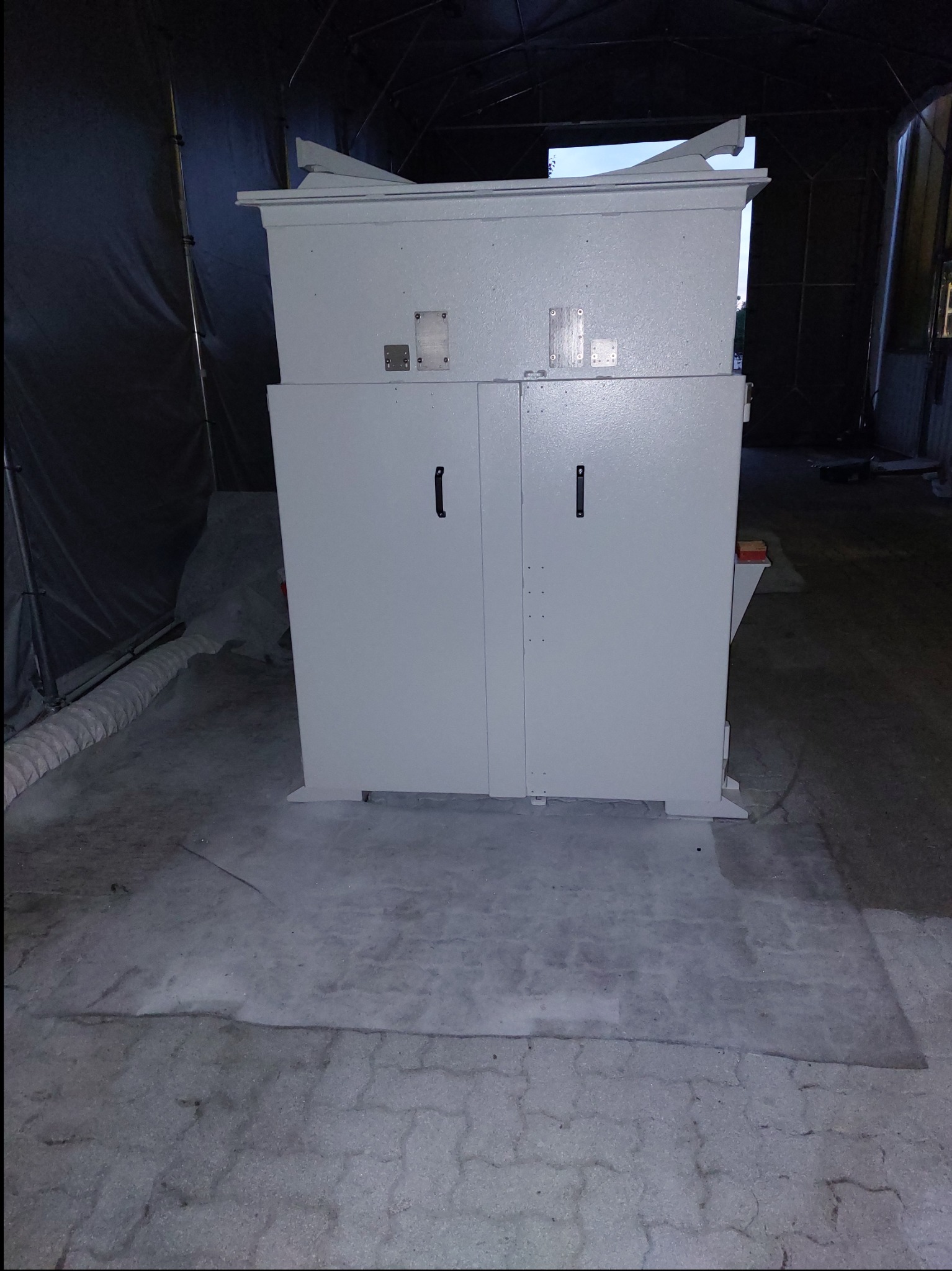 Biała, metalowa szafa z dwoma drzwiami i czarnymi uchwytami, umieszczona w pomieszczeniu o betonowej podłodze i ciemnym tle.