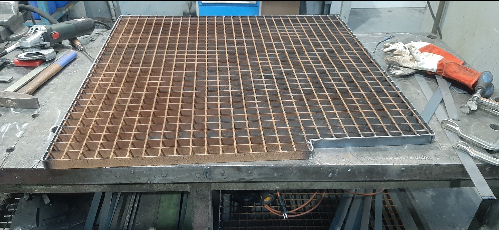 Metalowa krata z kwadratowymi oczkami, częściowo obramowana, leży na stole warsztatowym, obok narzędzia i rękawice robocze. Widoczny spaw na obramowaniu.