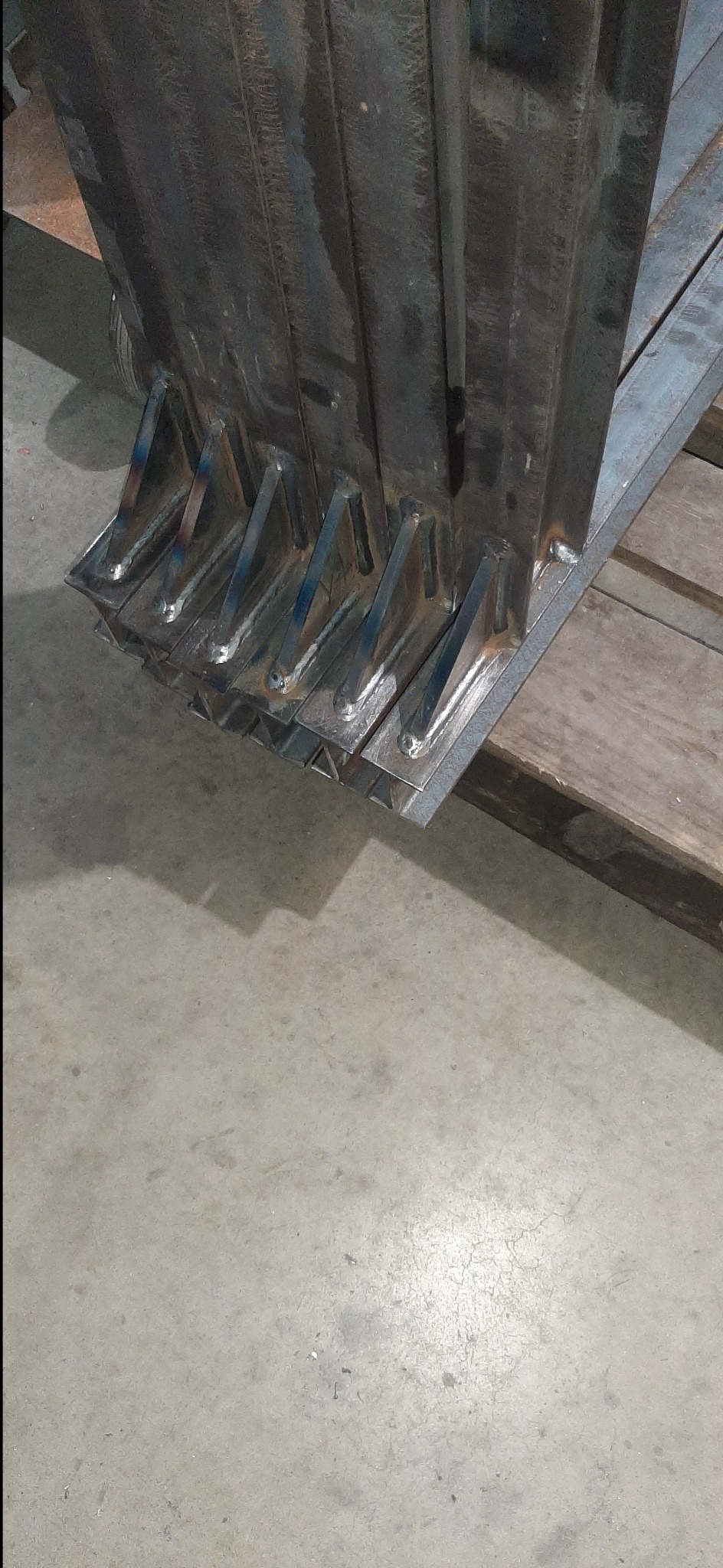 Surowe, spawane elementy konstrukcji metalowej z widocznymi śladami spawania, leżące na drewnianej palecie. Szara, betonowa posadzka w tle.