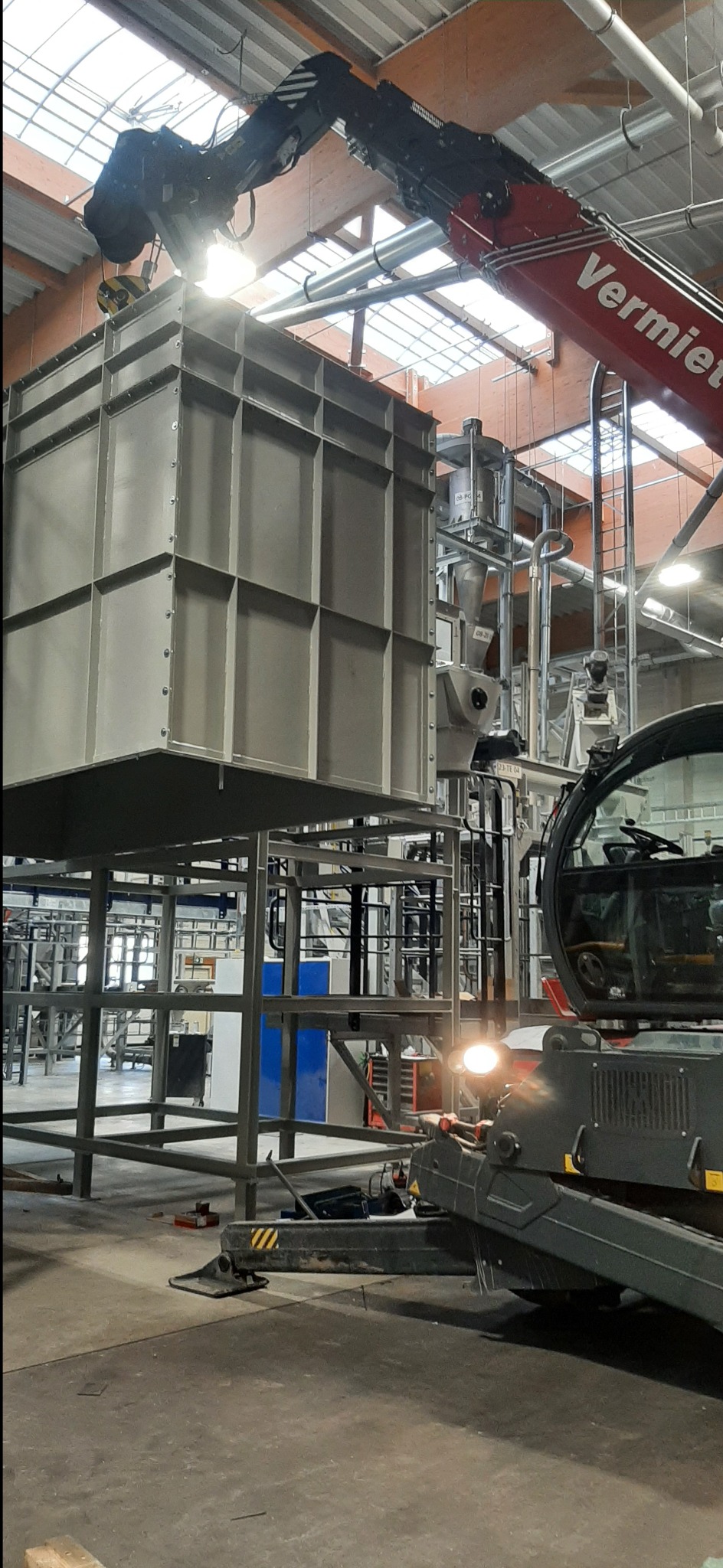 Klatka ze spawanych metalowych profili podnoszona przez dźwig w hali produkcyjnej. Widoczna konstrukcja stalowa, elementy instalacji przemysłowej i fragment kabiny dźwigu.