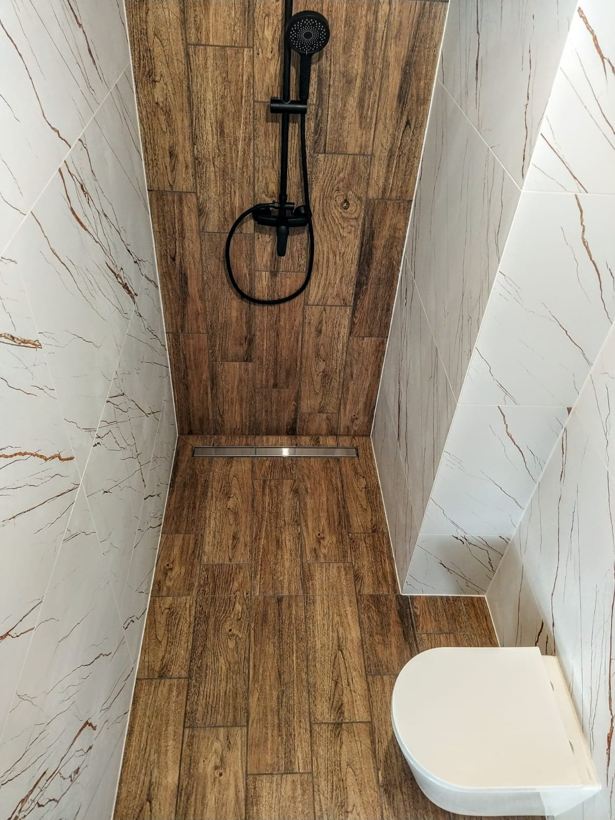 Nowoczesna kabina prysznicowa walk-in z odpływem liniowym, wyłożona płytkami imitującymi drewno w odcieniach brązu i białymi płytkami marmuropodobnymi z pomarańczowymi żyłkami, widoczny również...