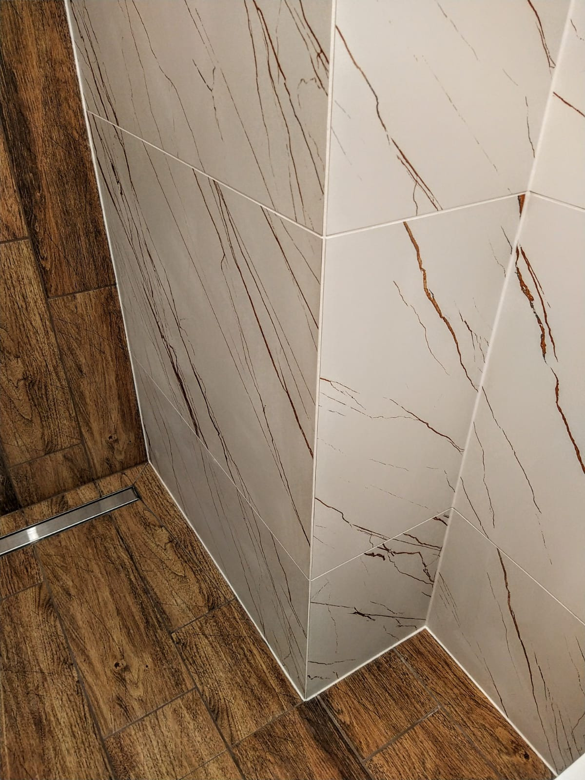 Kąt łazienki wykończony płytkami imitującymi drewno na podłodze i marmur na ścianach, z widocznym odpływem liniowym.