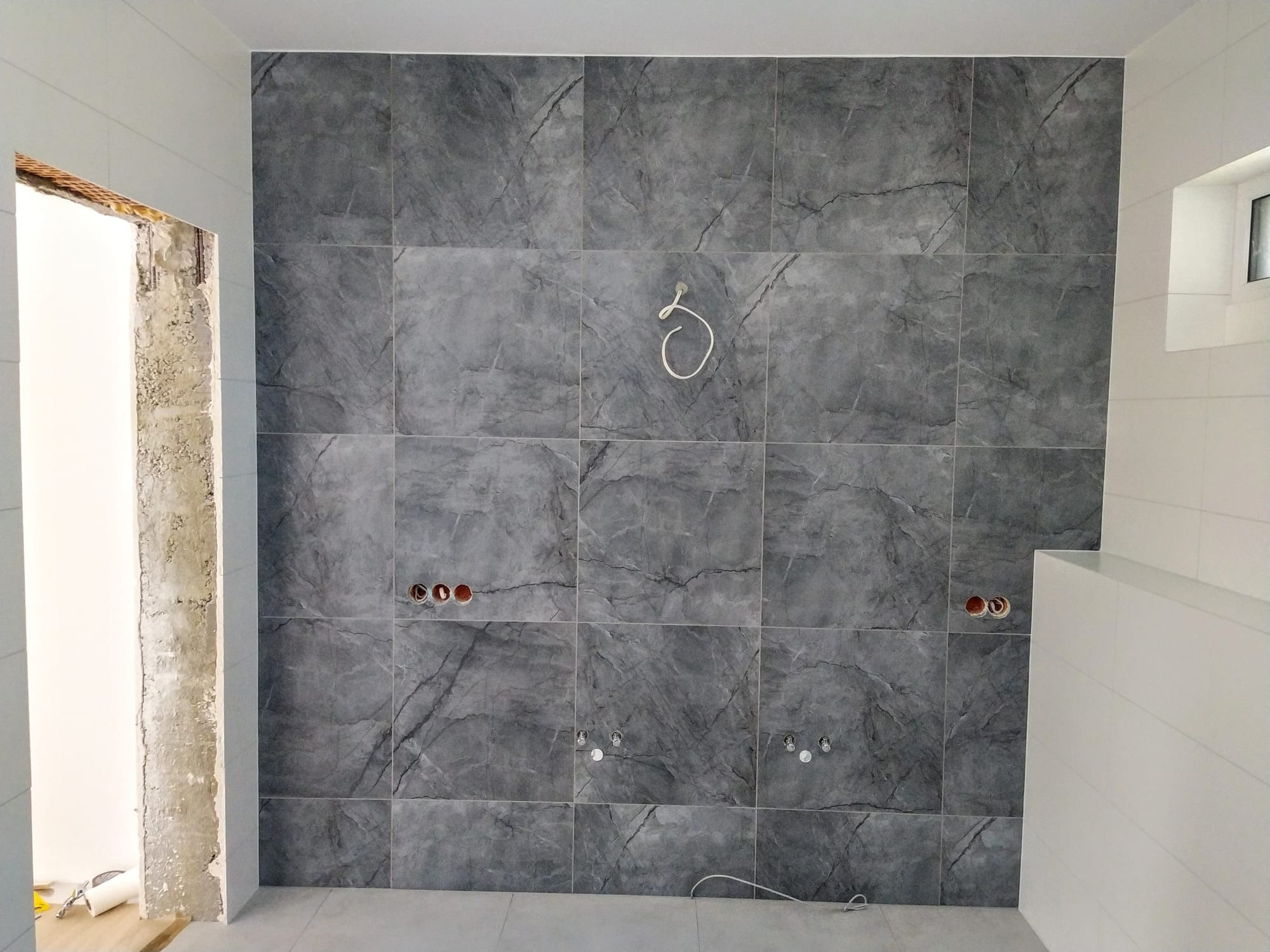 Ściana łazienki wyłożona szarymi płytkami imitującymi marmur, z widocznymi otworami na instalacje i wystającym przewodem elektrycznym; fragment surowego muru w otworze drzwiowym.