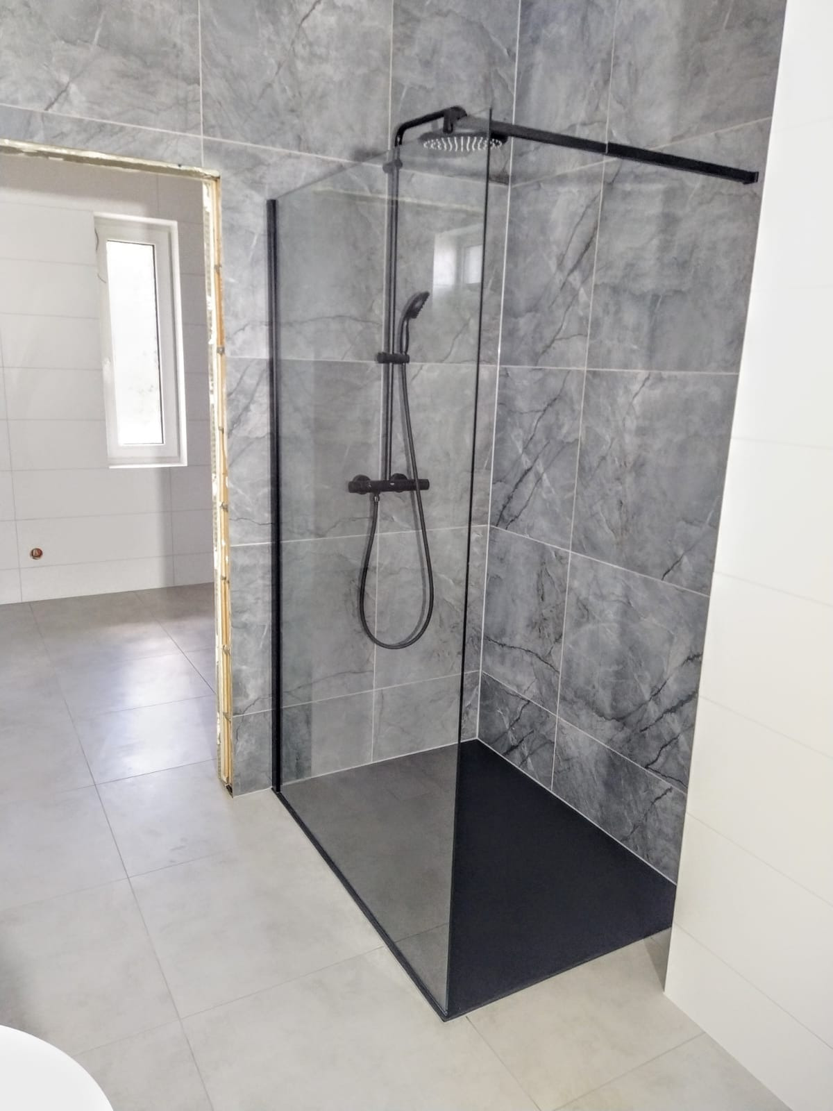Nowoczesna kabina prysznicowa typu walk-in z czarną armaturą i ciemnym brodzikiem, obłożona szarymi płytkami imitującymi marmur, widok na sąsiednie pomieszczenie z oknem.