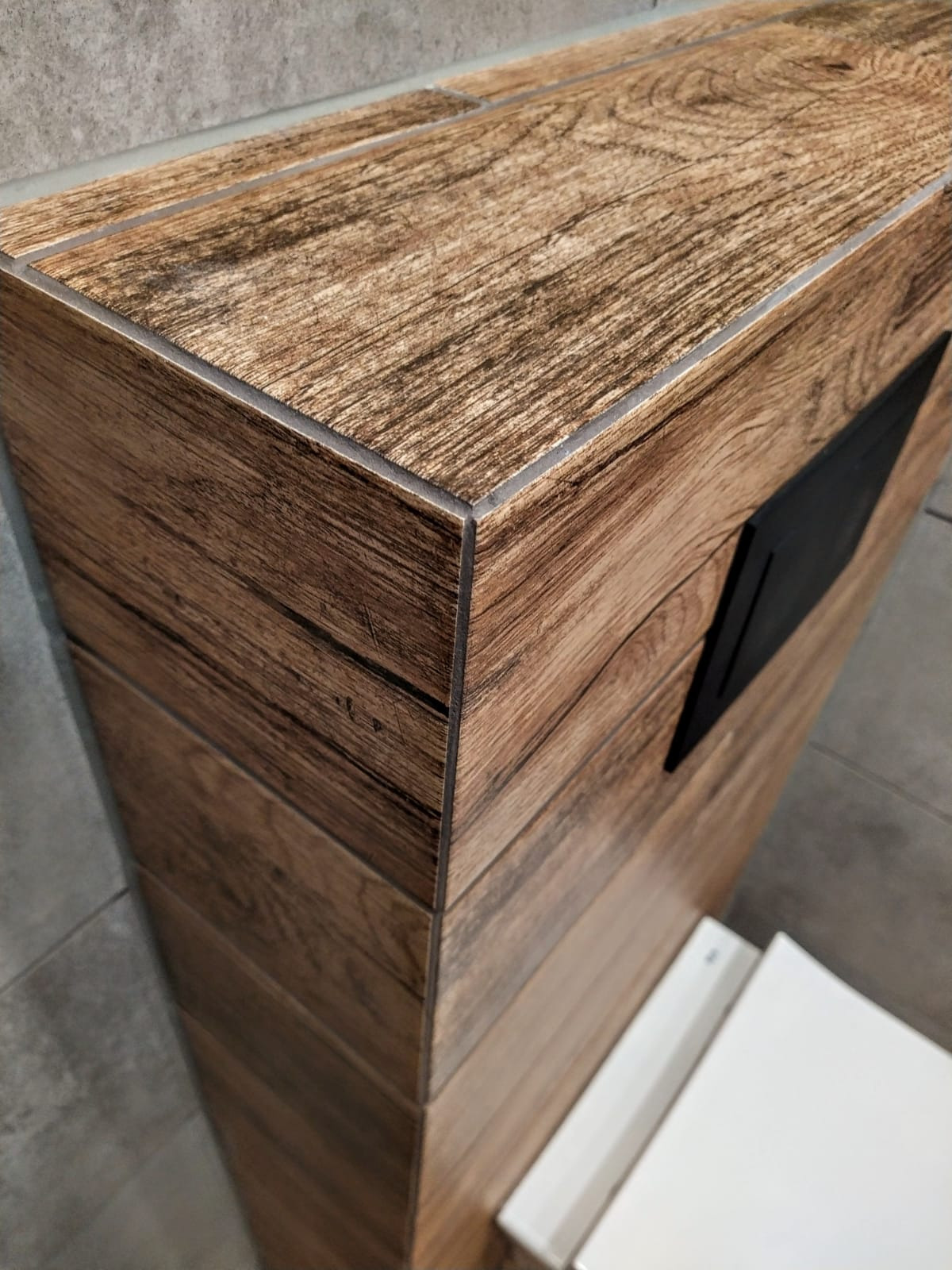 Wykończona obudowa z płytek imitujących drewno, z czarnym otworem rewizyjnym, szare płytki podłogowe, widok z góry.