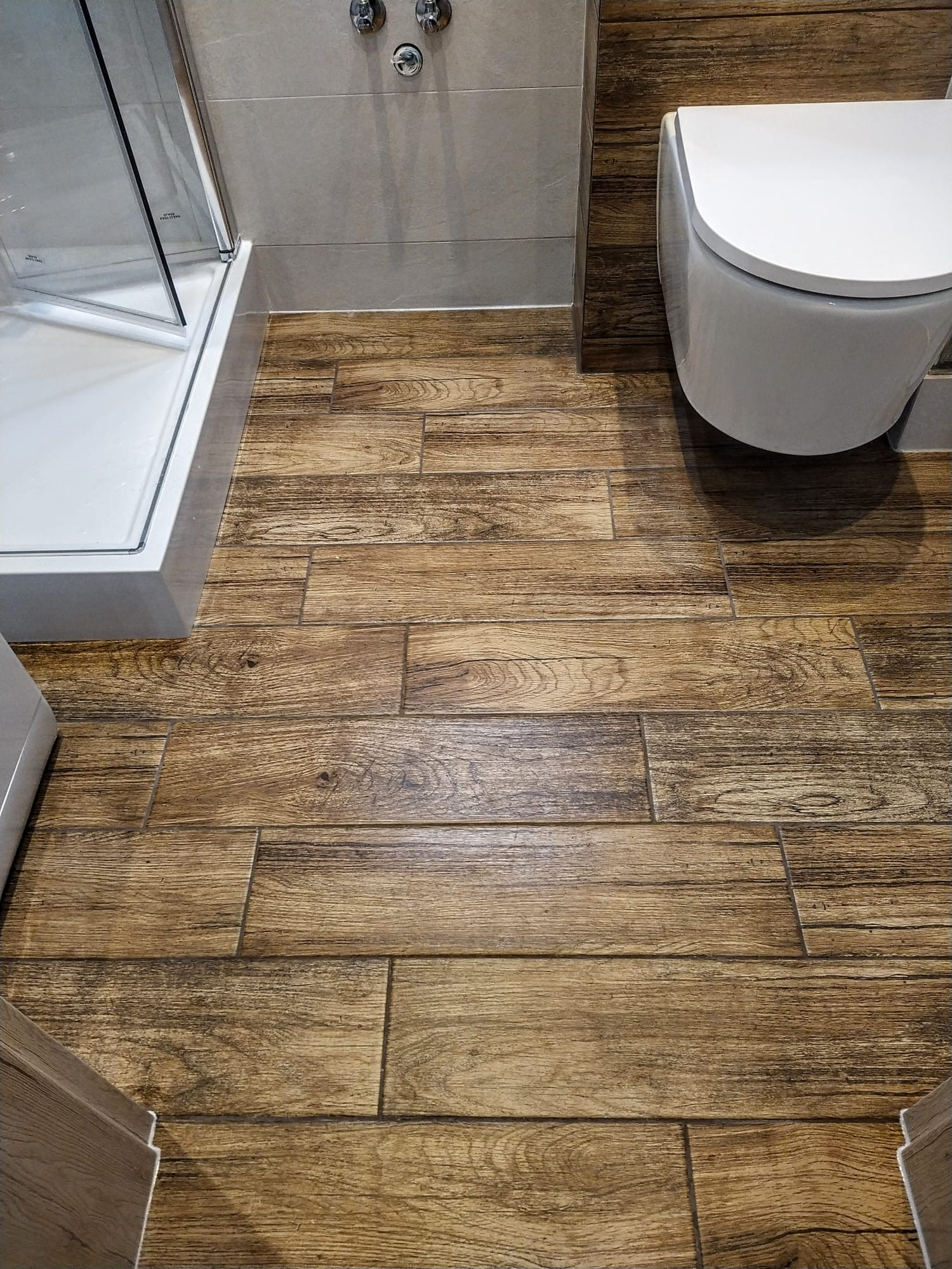 Ukończona łazienka z drewnopodobną podłogą, podwieszaną toaletą i kabiną prysznicową ze srebrnymi okuciami.