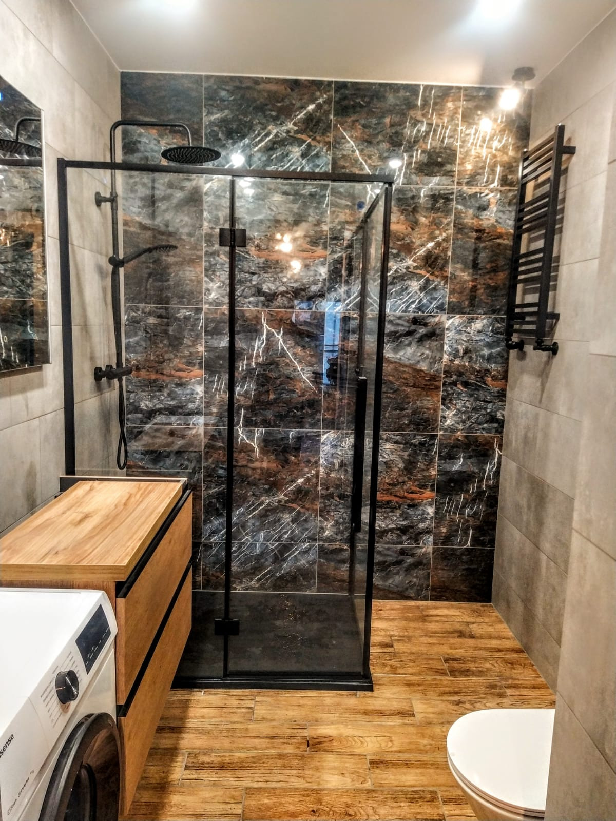 Nowoczesna łazienka z kabiną prysznicową typu walk-in z czarnymi profilami, płytki imitujące marmur na ścianie, drewnopodobna podłoga, pralka z drewnianą zabudową i grzejnik drabinkowy.