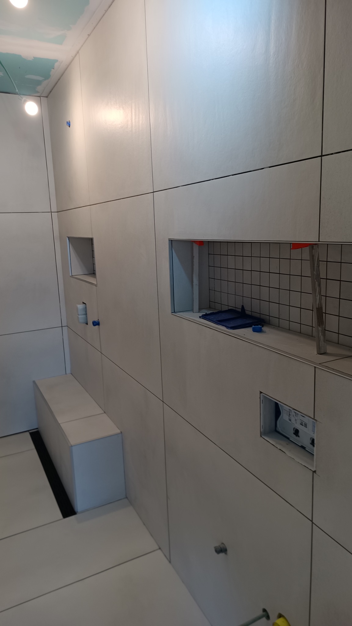 Ściana łazienki wyłożona wielkoformatowymi płytkami z wnękami, widoczne narzędzia glazurnicze i poziomica, sufit częściowo wykończony płytami gipsowo-kartonowymi.