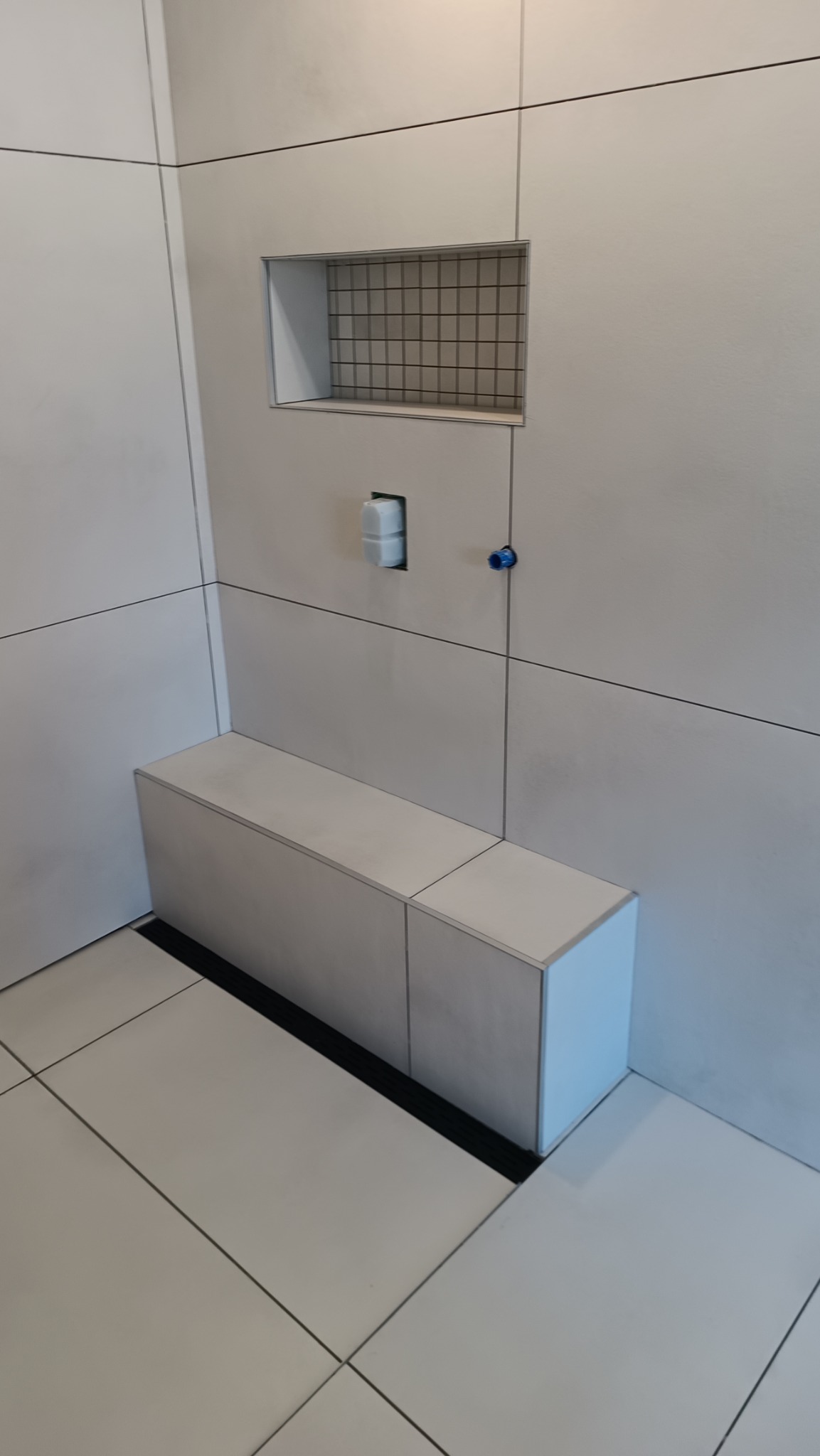 Nowoczesna łazienka z jasnymi, kwadratowymi płytkami, wnęką wyłożoną drobną mozaiką, czarnym odpływem liniowym i zabudową z płytek imitującą siedzisko lub półkę.