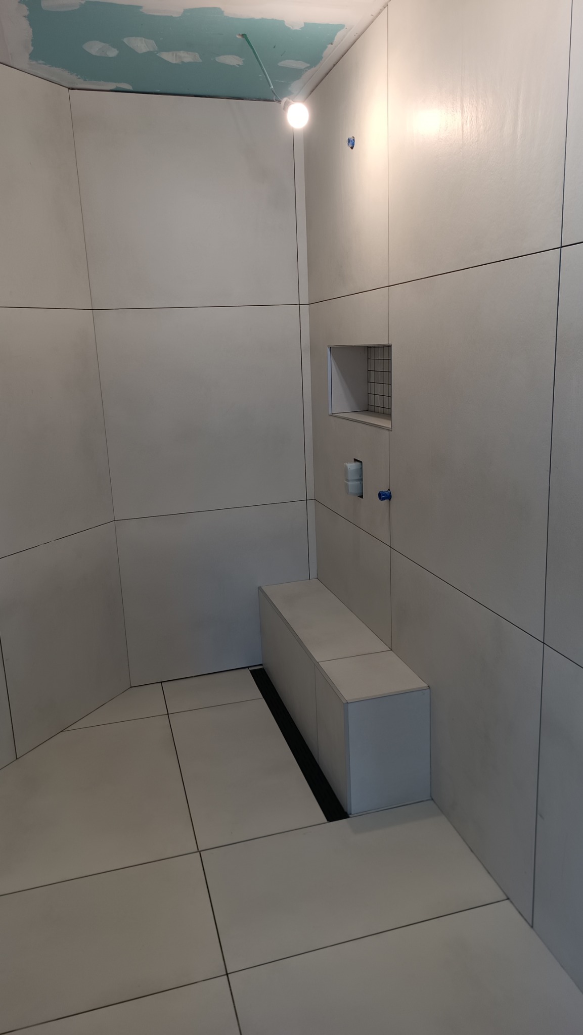 Nowoczesna łazienka w trakcie wykańczania: białe, wielkoformatowe płytki, wnęka z siatką, podwieszana półka/siedzisko z odpływem liniowym, surowy sufit z widocznymi śladami gładzi.