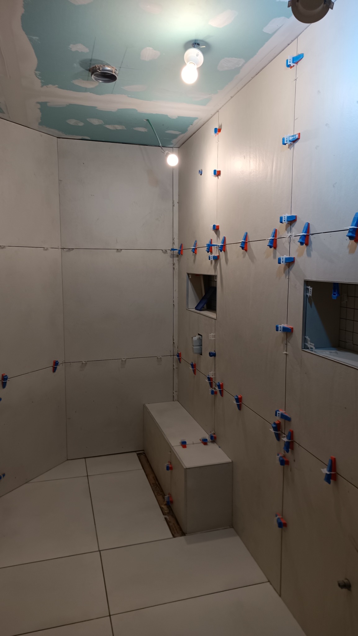 Łazienka w trakcie układania płytek, widoczne poziomice laserowe i klipsy do płytek na ścianach, sufit w stanie surowym z widocznymi elementami instalacji, podłoga wyłożona jasnymi płytkami