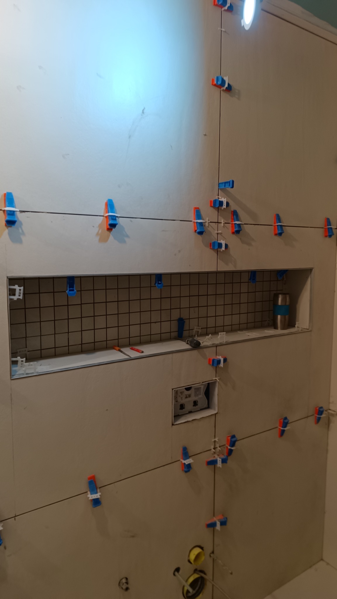 Ściana w trakcie układania płytek, z widocznymi klipsami poziomującymi i wnęką z metalową siatką, oświetlona punktowym światłem.