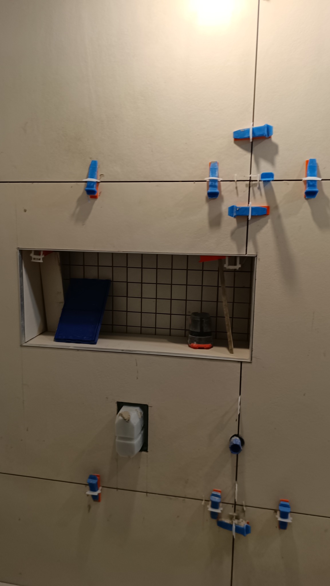 Ściana z jasnych płytek w trakcie montażu, widoczne niebiesko-pomarańczowe klipsy poziomujące, wnęka z kratką, niebieskie płytki i narzędzia w środku, otwór na instalację.