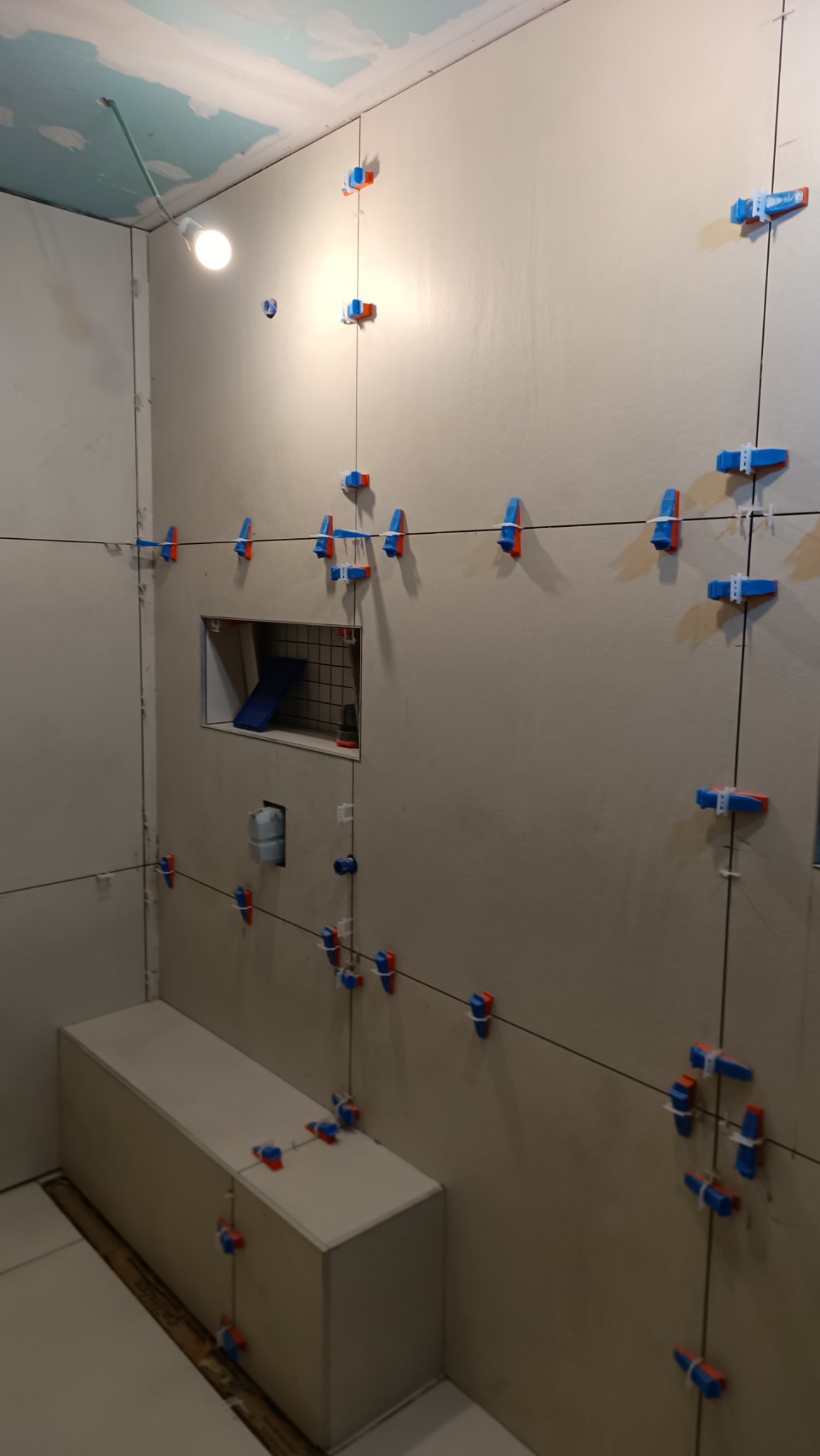 Ściana łazienki w trakcie układania płytek, widoczne listwy poziomujące, niebiesko-pomarańczowe klipsy, wnęka w ścianie, surowy sufit i prowizoryczne oświetlenie.