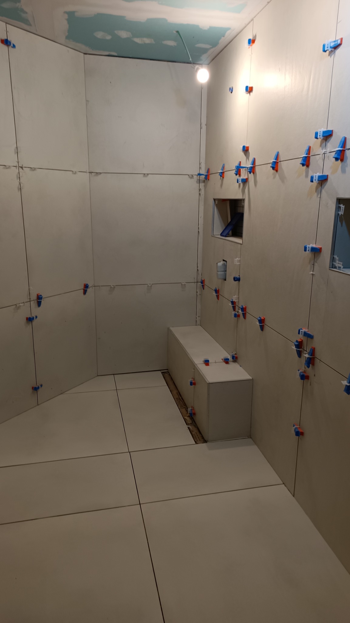 Białe płytki podłogowe i ścienne w trakcie montażu w łazience, z widocznymi systemami poziomowania płytek, surowy sufit w kolorze niebieskim, wnęka w ścianie.