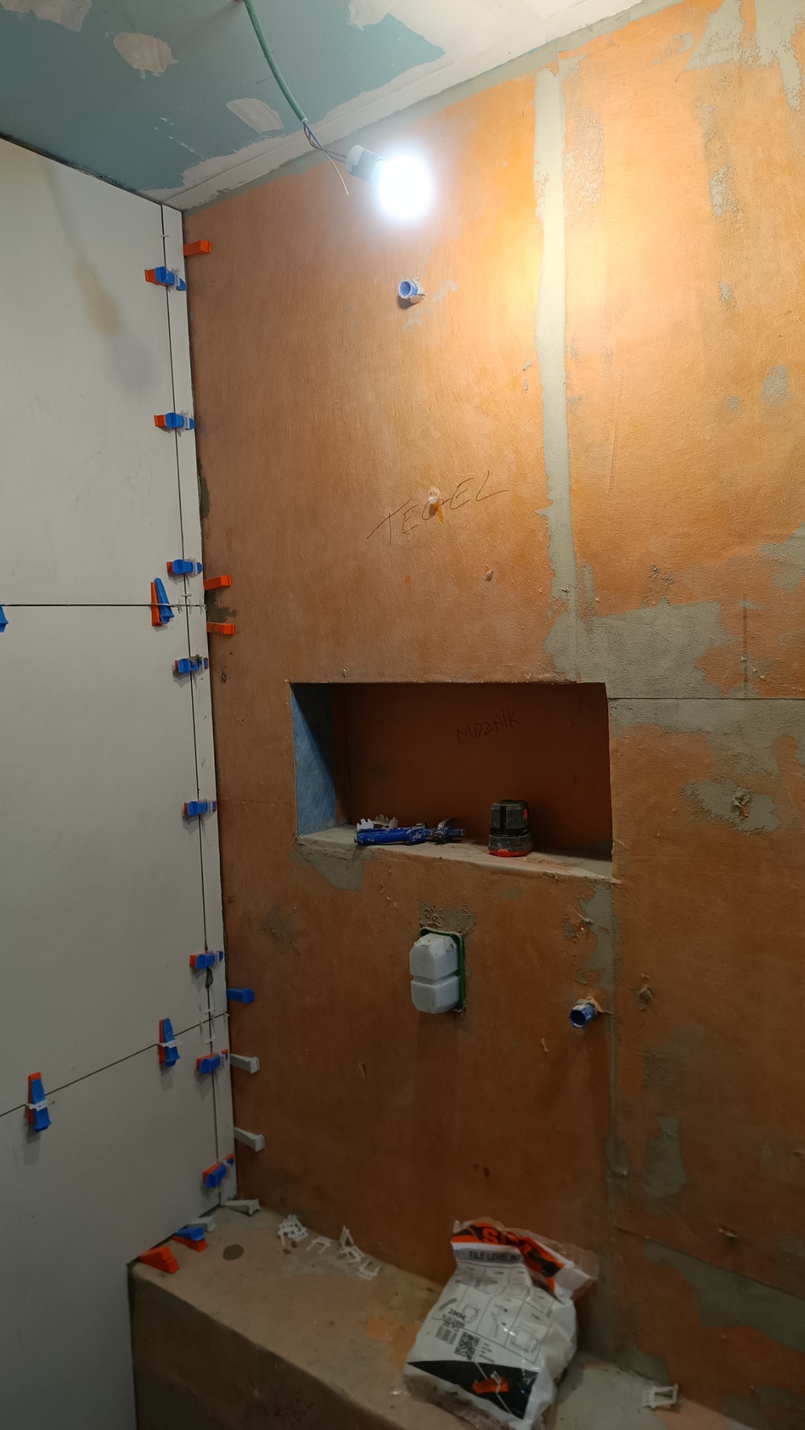 Ściana łazienki w trakcie układania płytek, widoczne krzyżyki poziomujące, wnęka w ścianie z zapisanym słowem 'Mozaik' oraz laserowy niwelator, ściana zagruntowana na pomarańczowo z napisem 'Tegel'.