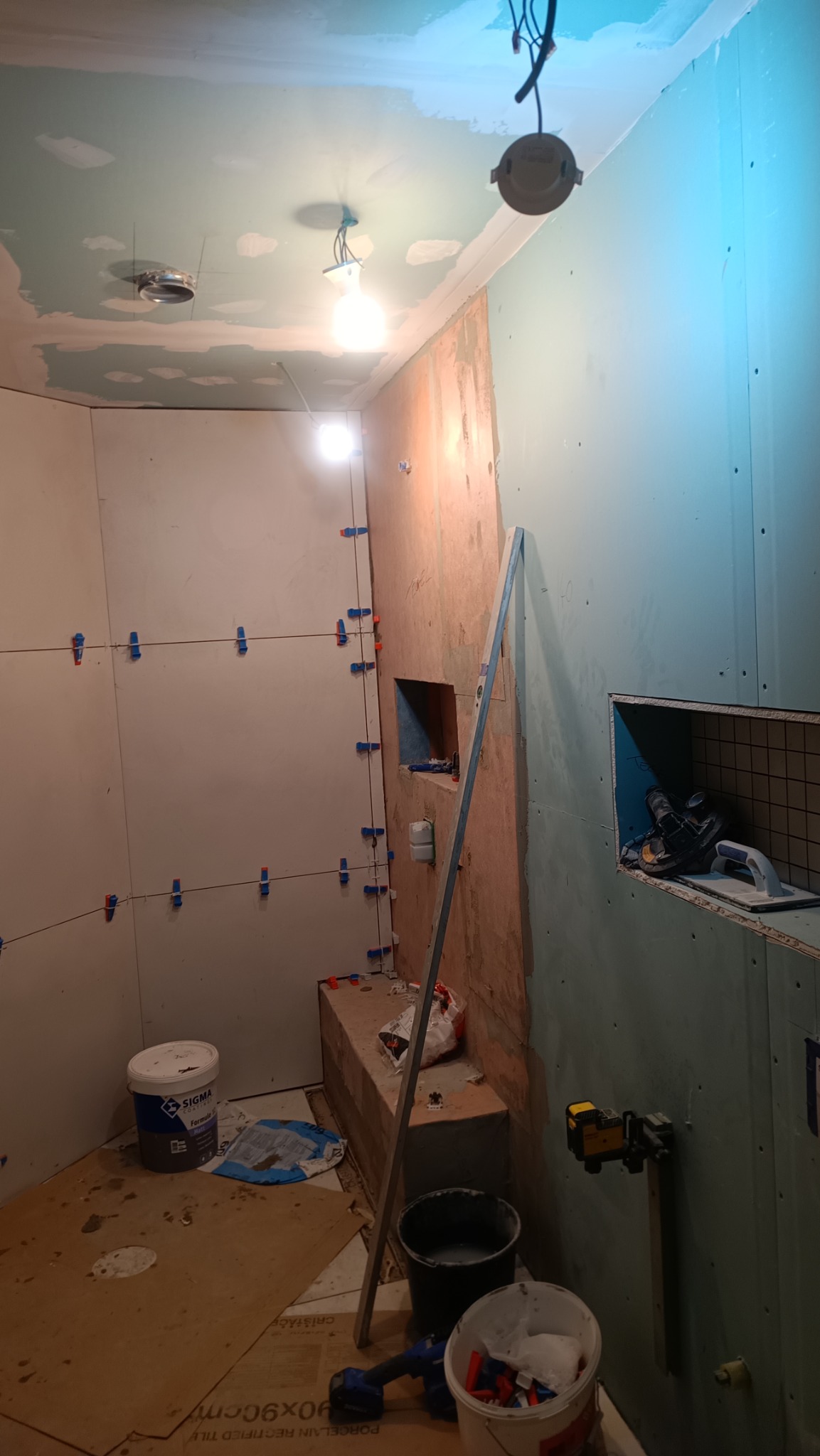 Łazienka w trakcie remontu: ściany przygotowane pod glazurę, widoczne listwy poziomujące, narożniki z klipsami, na podłodze płyty kartonowo-gipsowe, narzędzia i wiadro z klejem.