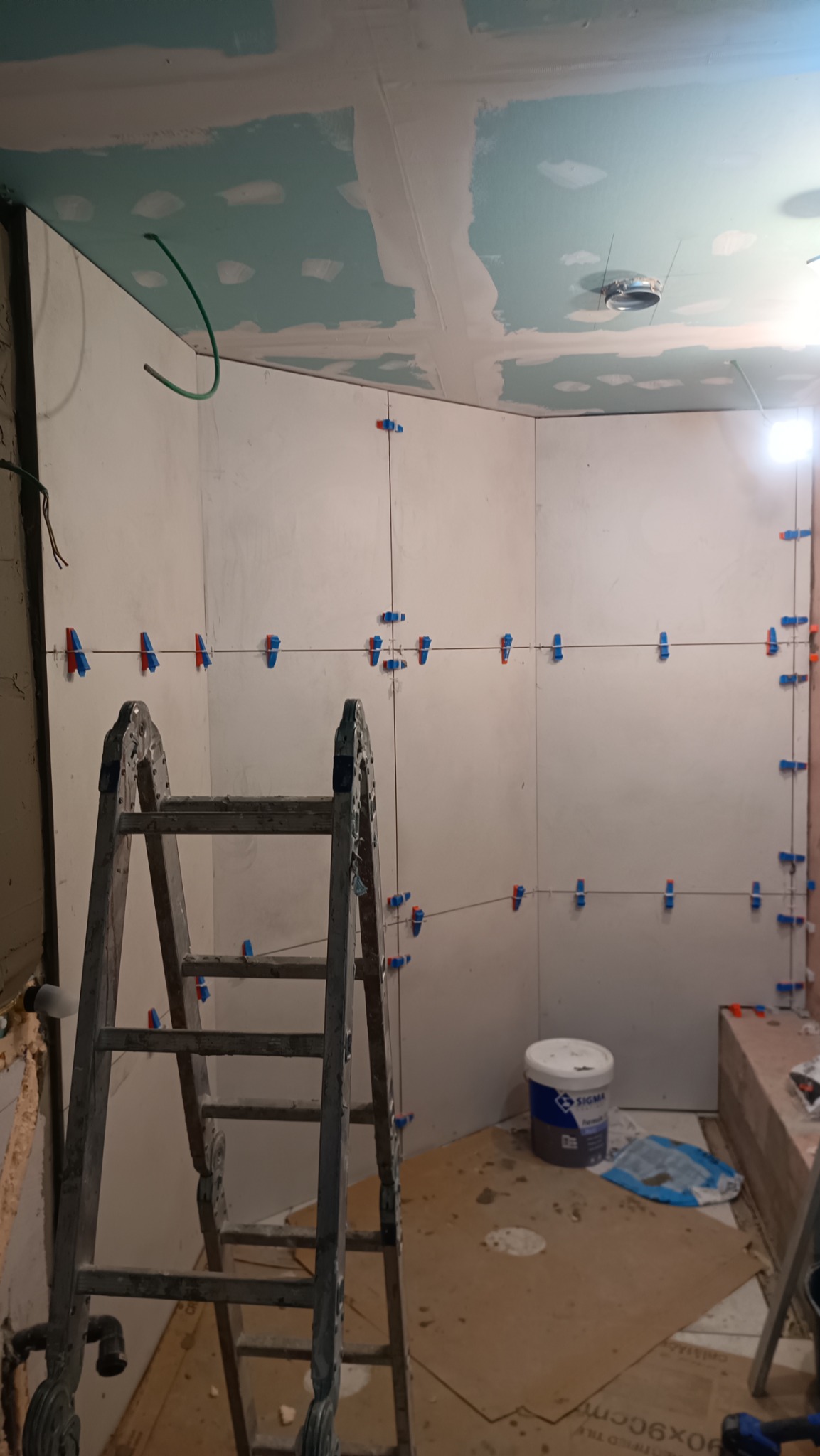 Przygotowanie do układania płytek w łazience, widoczne wypoziomowane ściany z zamocowanymi klipsami dystansowymi, drabina i wiadro z klejem.