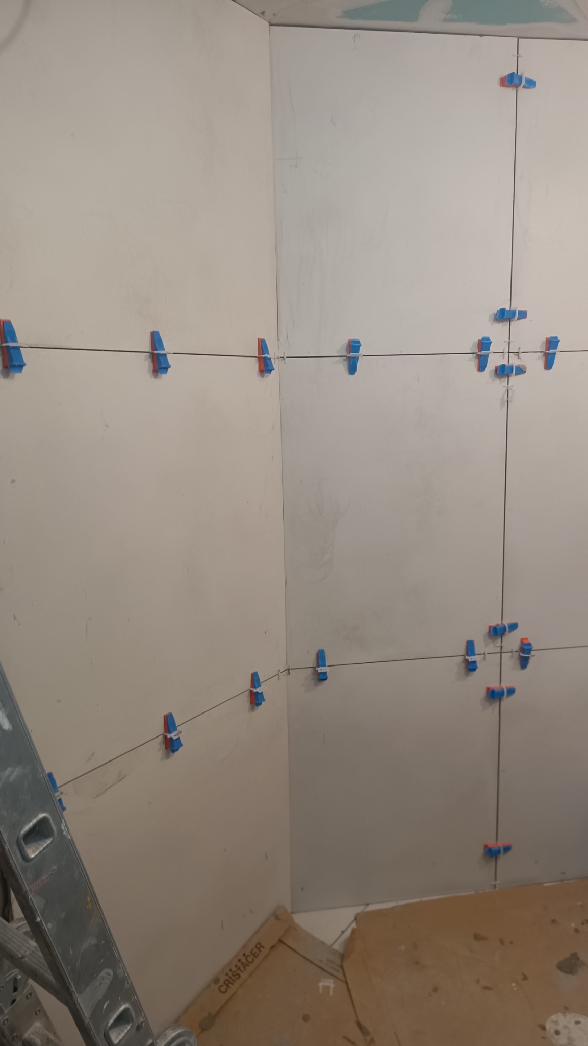 Świeżo ułożone białe płytki w rogu pomieszczenia, z użyciem poziomujących klipsów i sznurków, widoczne ślady zaprawy i drabina.