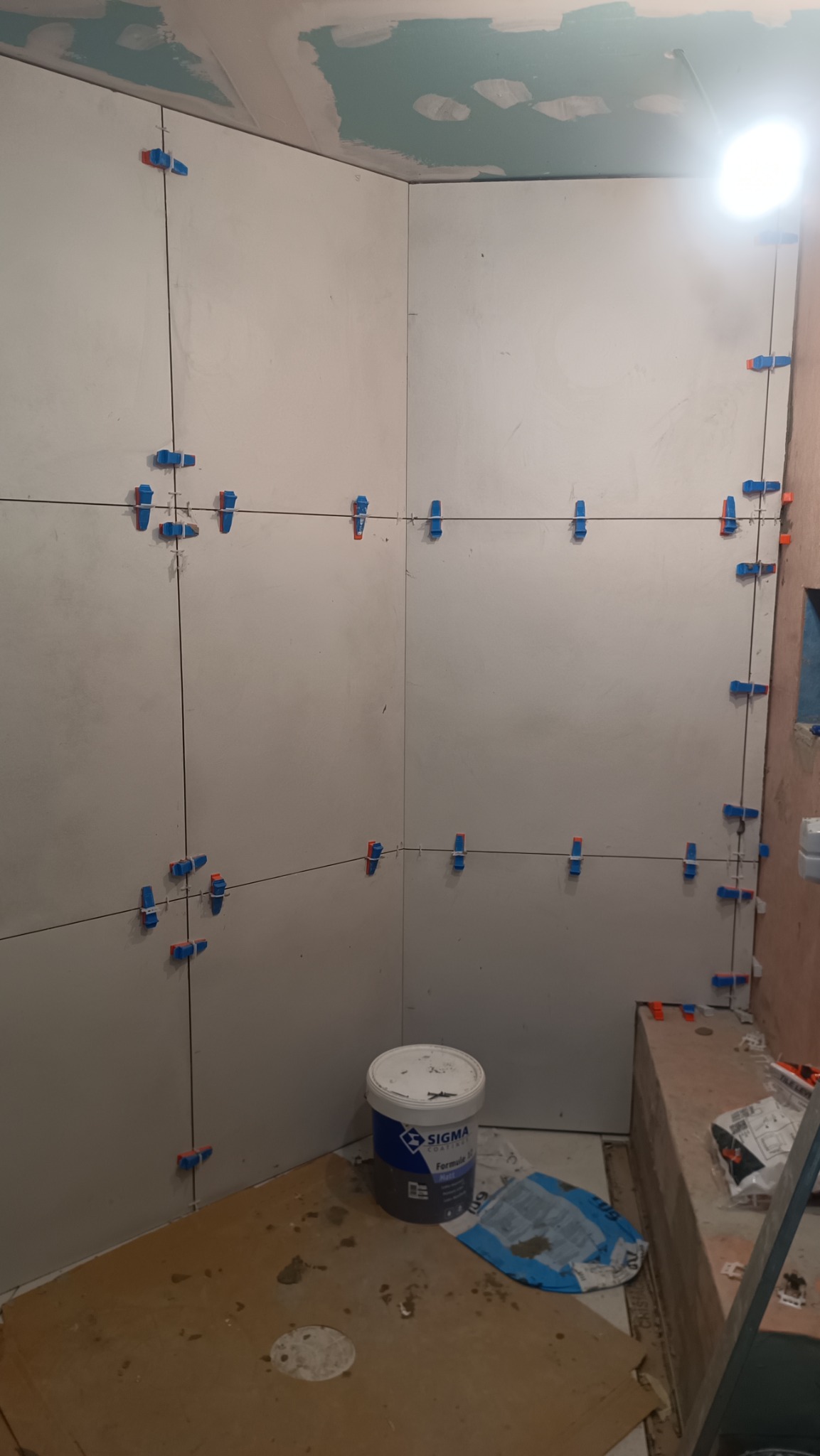 Przygotowanie do układania płytek w narożniku łazienki, z użyciem poziomych i pionowych linii oraz klipsów dystansowych, widoczny kubeł z zaprawą klejową Sigma i worek z klejem. Sufit w trakcie...