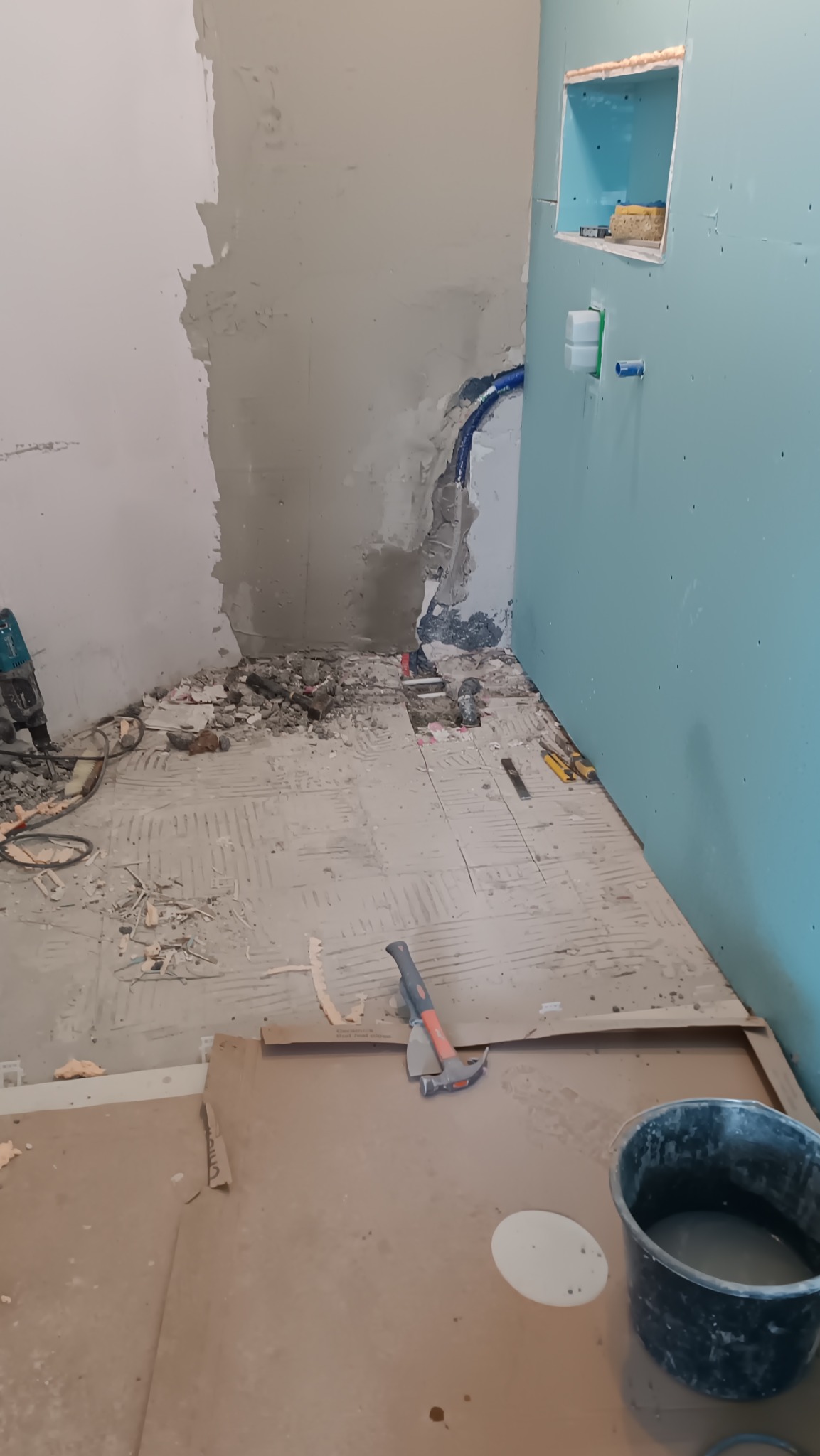 Demontaż starej posadzki w łazience, widoczne gruz, narzędzia, odkryte rury, ściany przygotowane pod nowe wykończenie, częściowo obłożone płytami gipsowo-kartonowymi z wnęką.